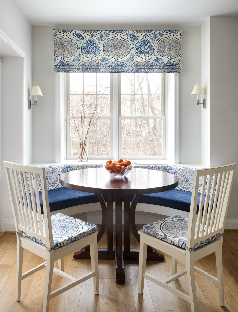 Breakfast nook with curved banquette, round dark wood table, white chairs, blue-and-white patterned fabric, and large window.