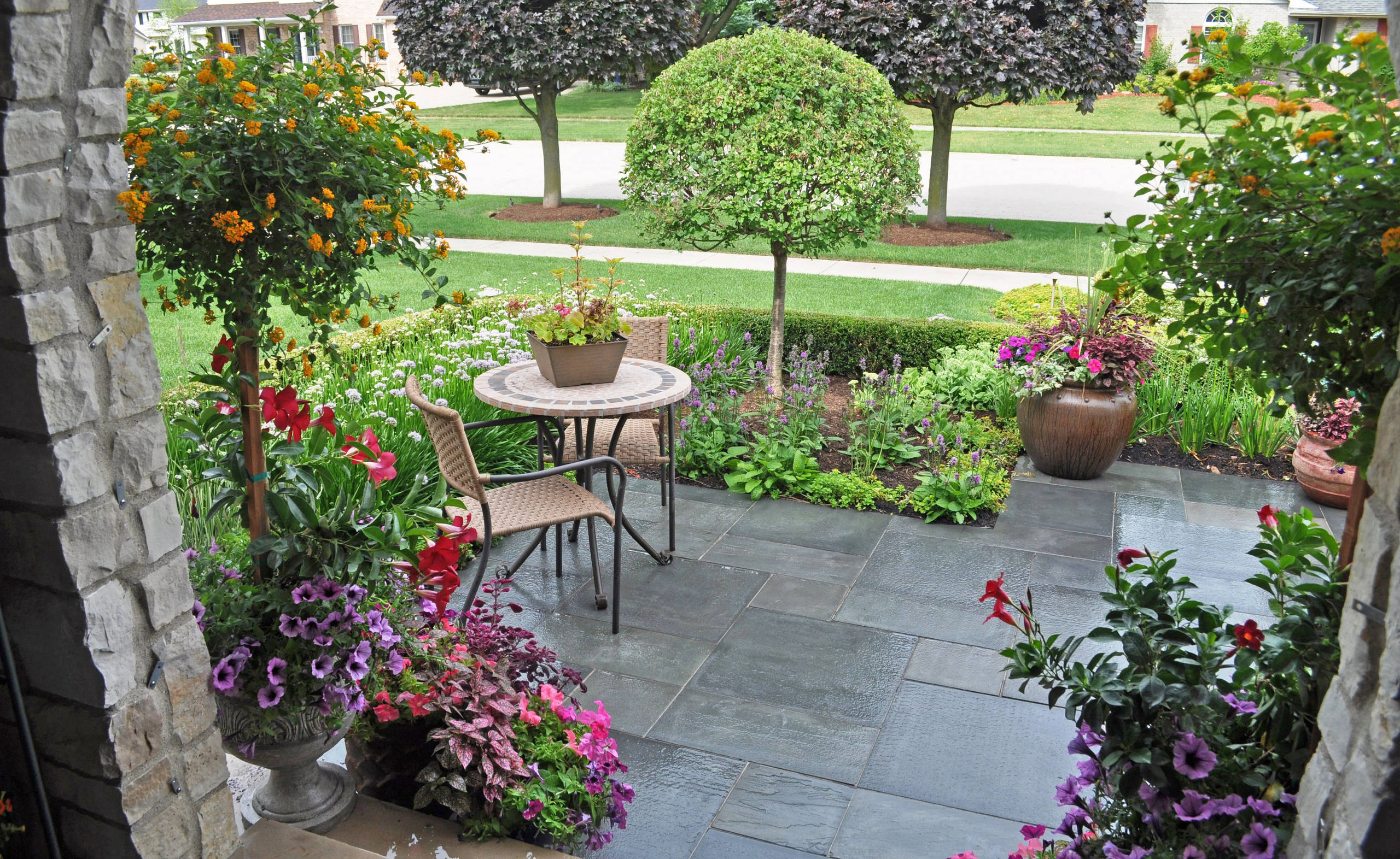 Bluestone patio with mosaic bistro table surrounded by colorful container gardens, manicured shrubs, and flowering perennials.
