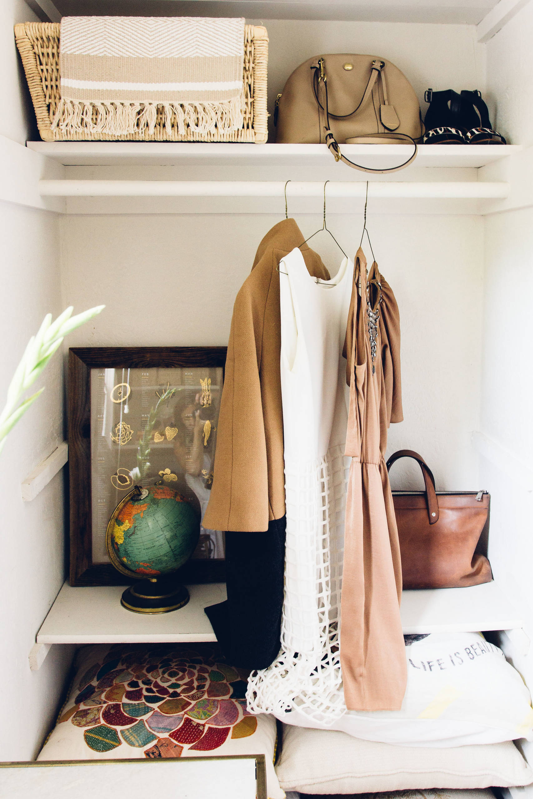 Compact closet alcove with hanging camel coat, woven basket, vintage globe, framed jewelry display, and leather accessories in neutral tones.