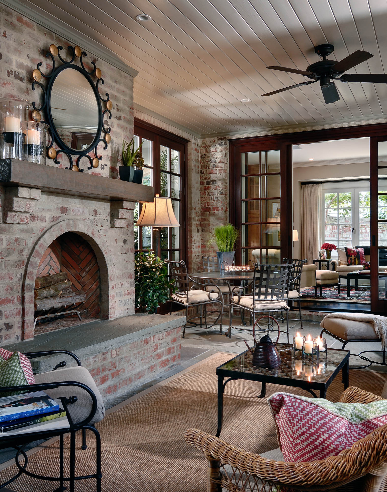 Sunroom with whitewashed brick walls, herringbone fireplace, ornate mirror, iron furniture, and sisal rug beneath paneled ceiling.