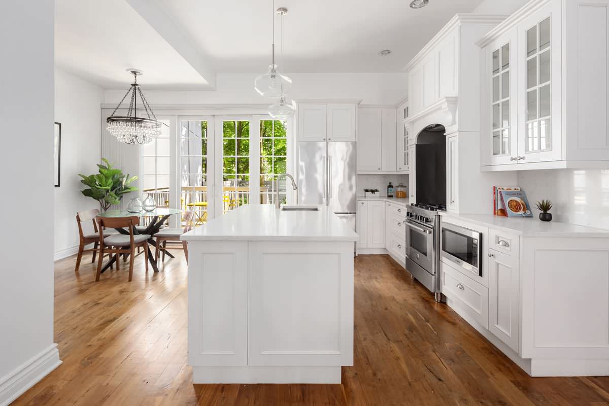 Open-concept white kitchen with marble island, crystal chandelier, French doors, hardwood floors, and adjacent dining area.