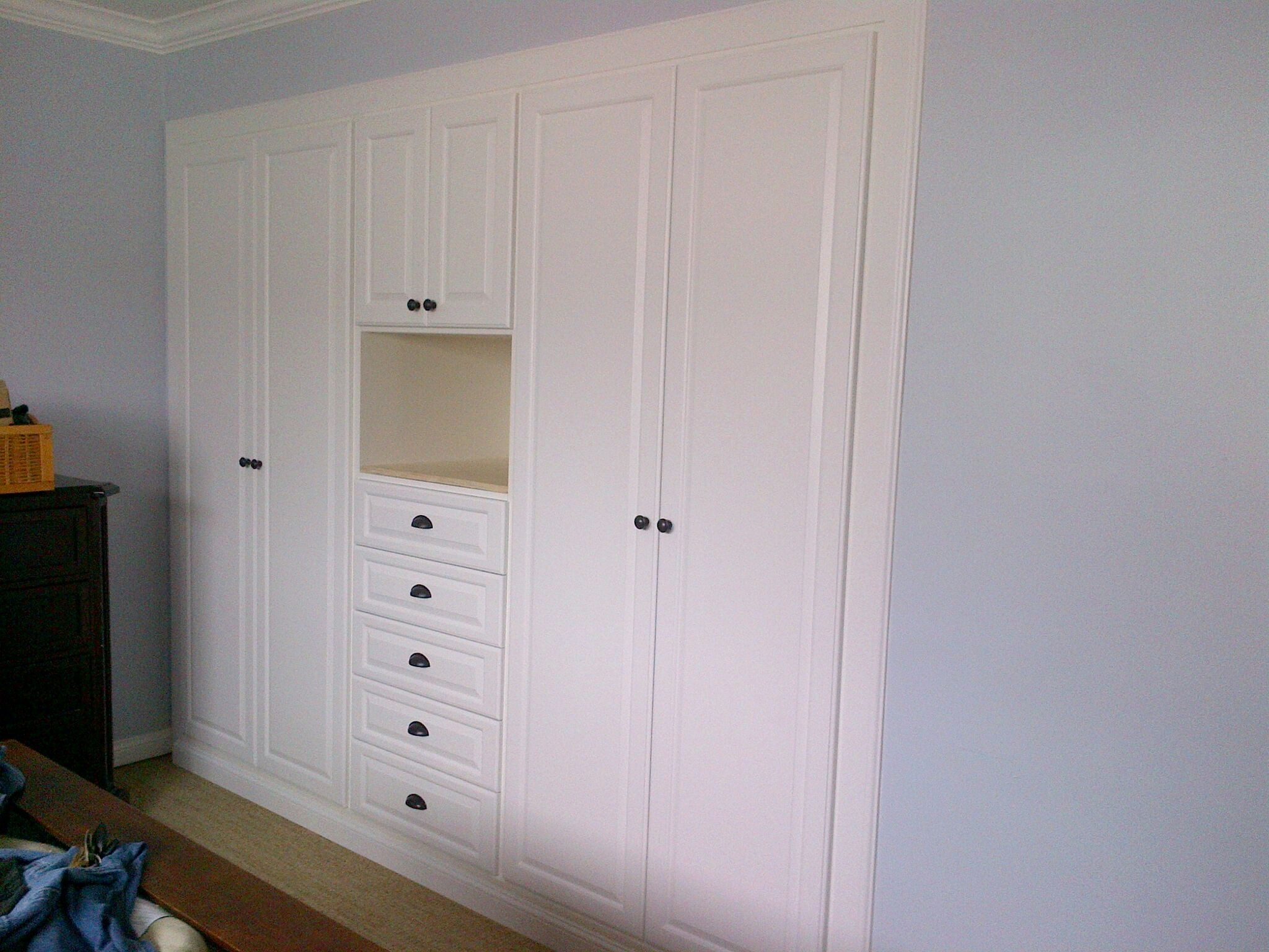Floor-to-ceiling white built-in wardrobe with central drawers, black hardware, and recessed nook against pale blue walls.