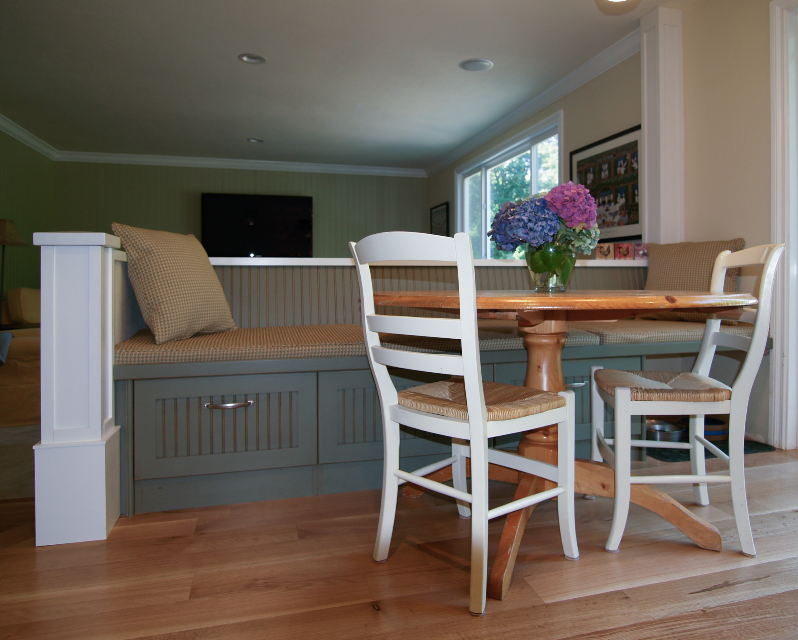 Breakfast nook featuring sage green built-in bench seating, white ladder-back chairs, round wooden table, and hydrangea centerpiece.