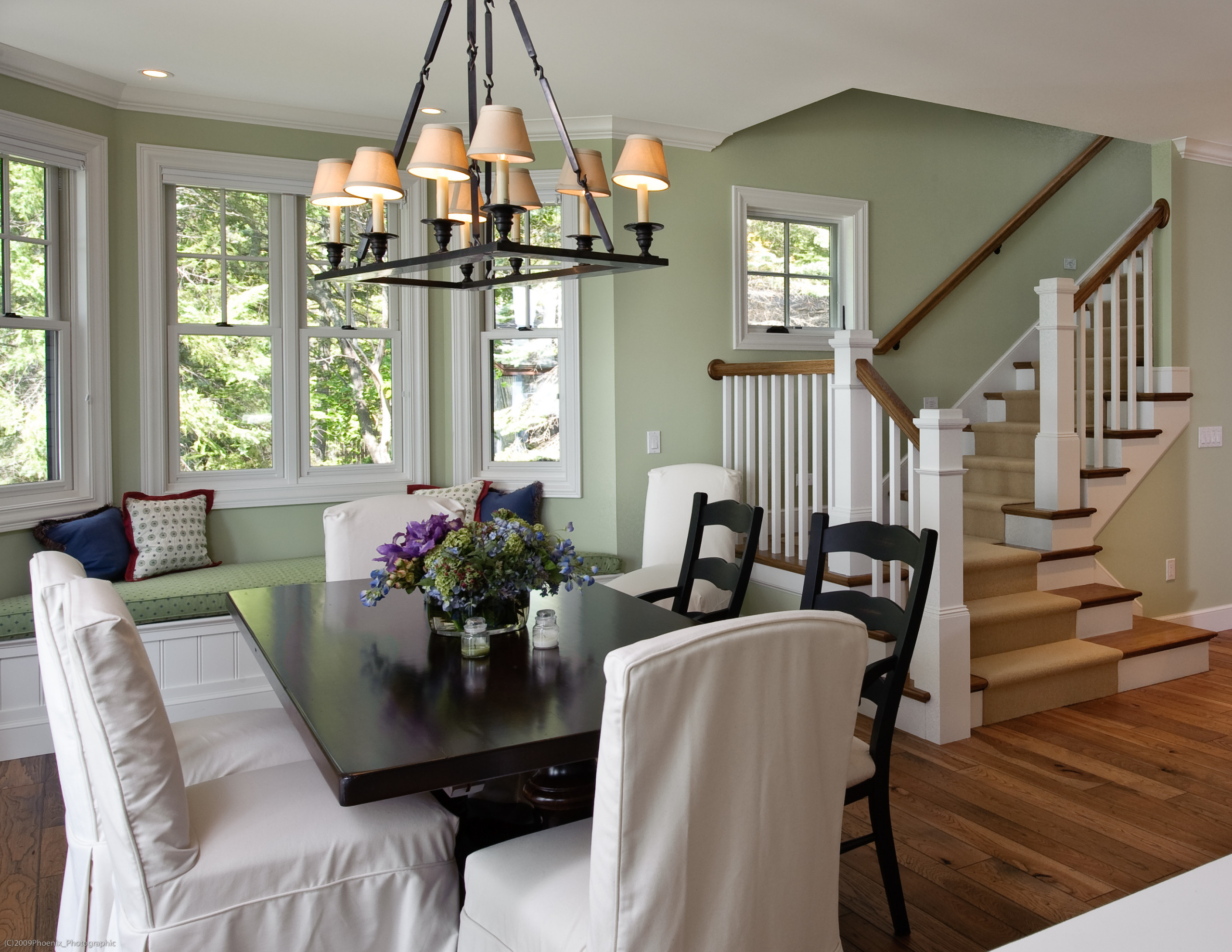 Dining room with built-in window bench, sage green walls, black dining table, white chairs, and iron chandelier.