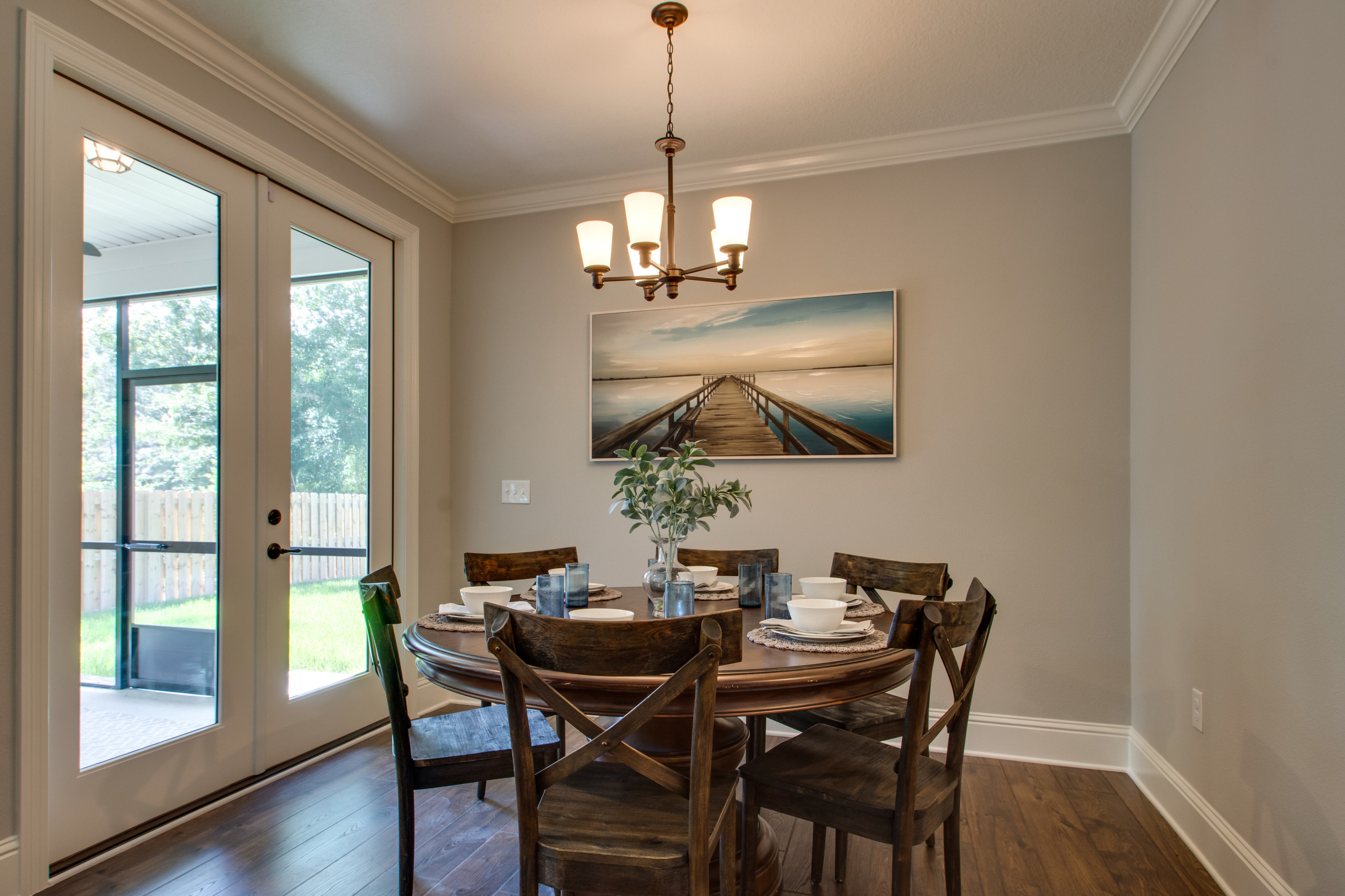 Dining room with round wooden table, X-back chairs, French doors, coastal artwork, and brass chandelier against greige walls.