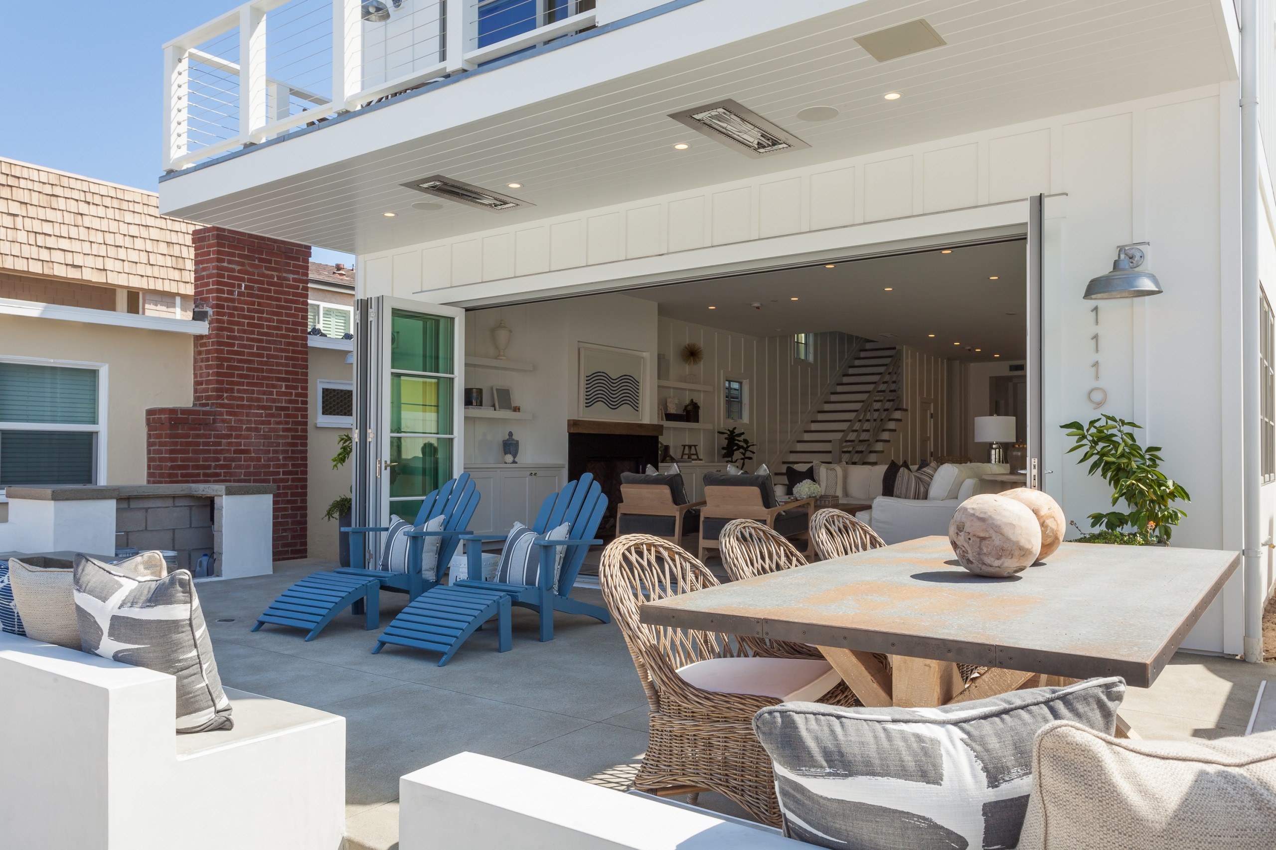 Indoor-outdoor living space with blue Adirondack chairs, wicker dining set, and folding glass doors opening to white coastal interior.