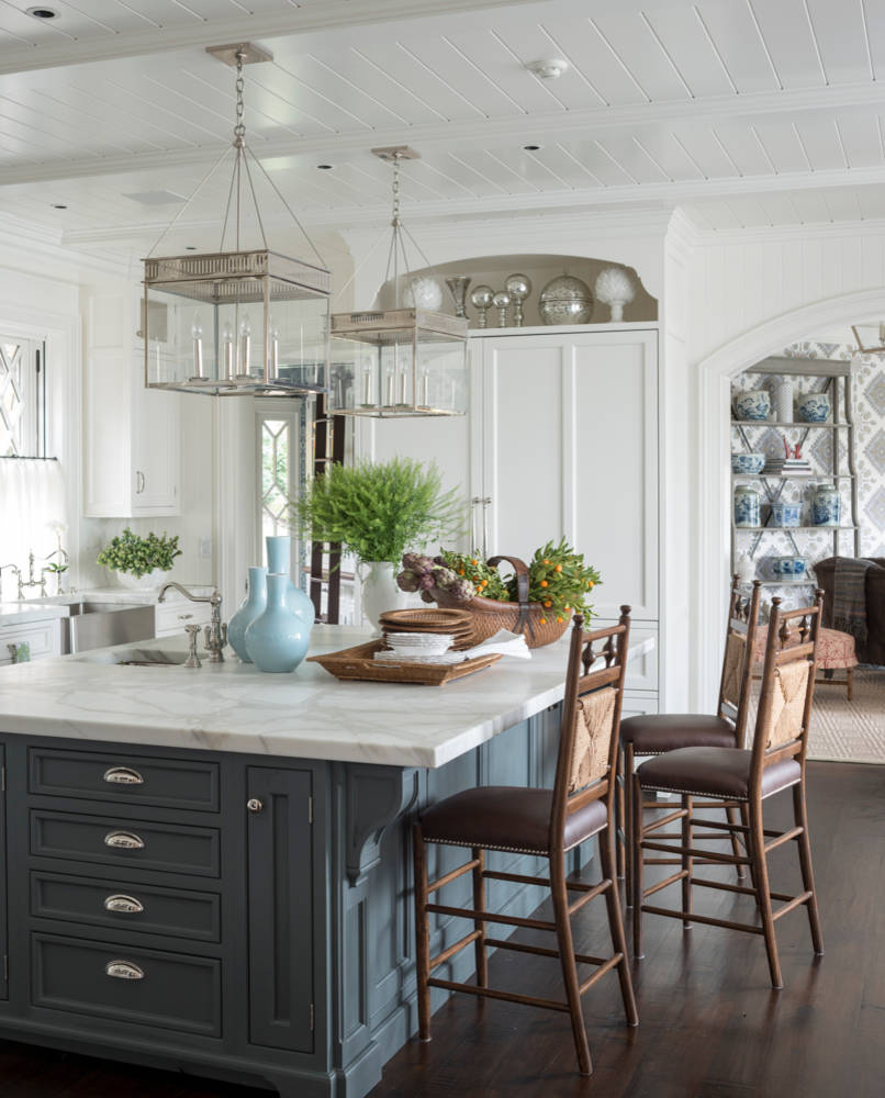 Bright kitchen with navy island, marble countertop, glass lantern pendants, and carved wooden barstools beneath planked ceiling.
