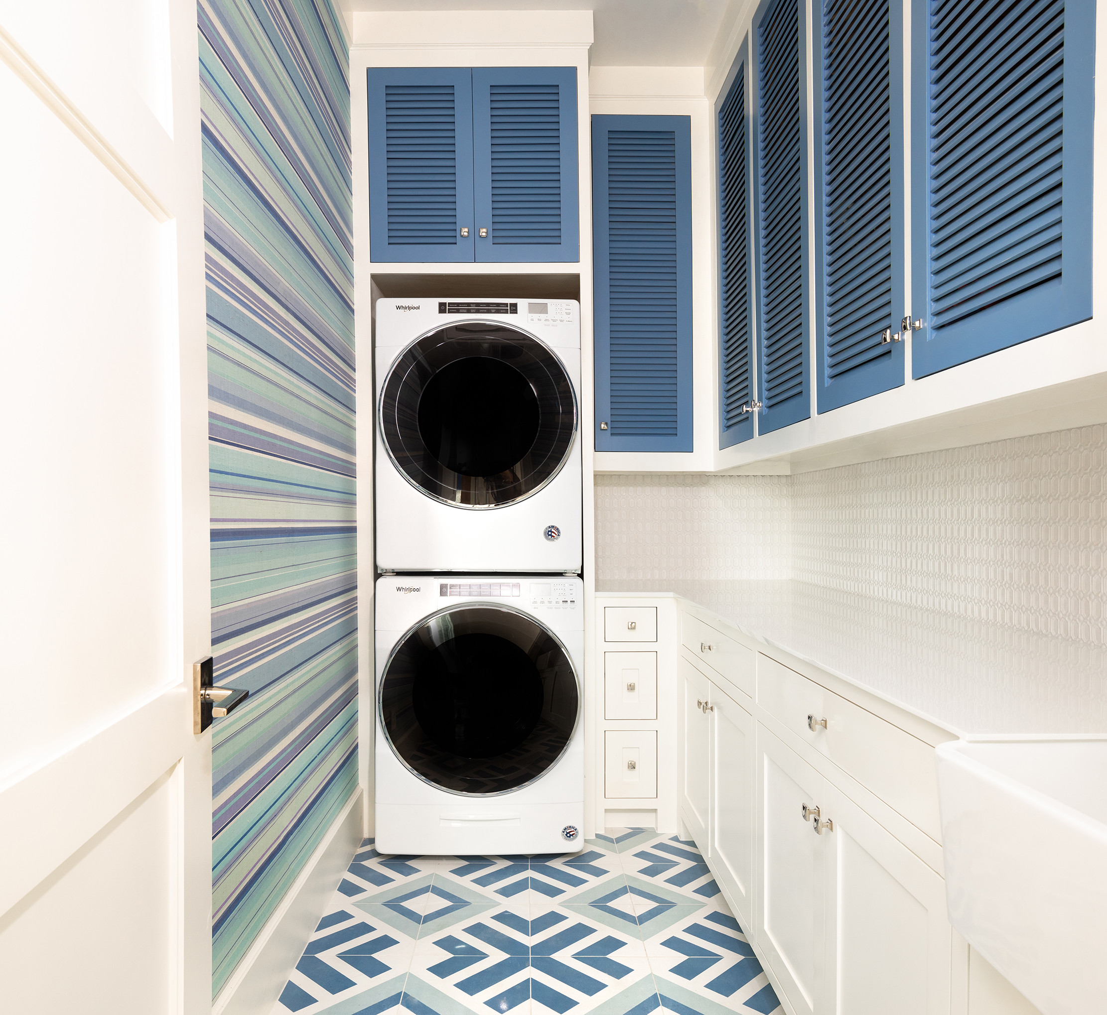 Modern laundry room with stacked white appliances, blue louvered cabinets, geometric floor tiles, and striped wallpaper.