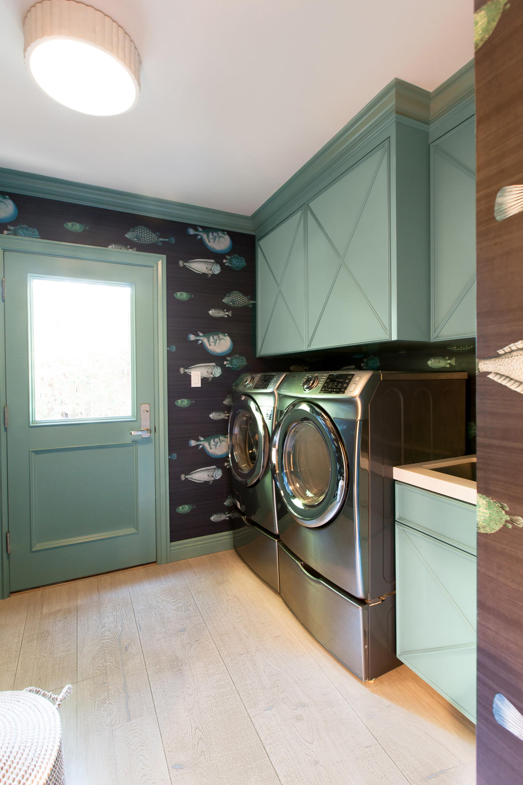 Laundry room with sage green cabinets, fish-patterned wallpaper, stainless steel appliances, and light wood flooring.
