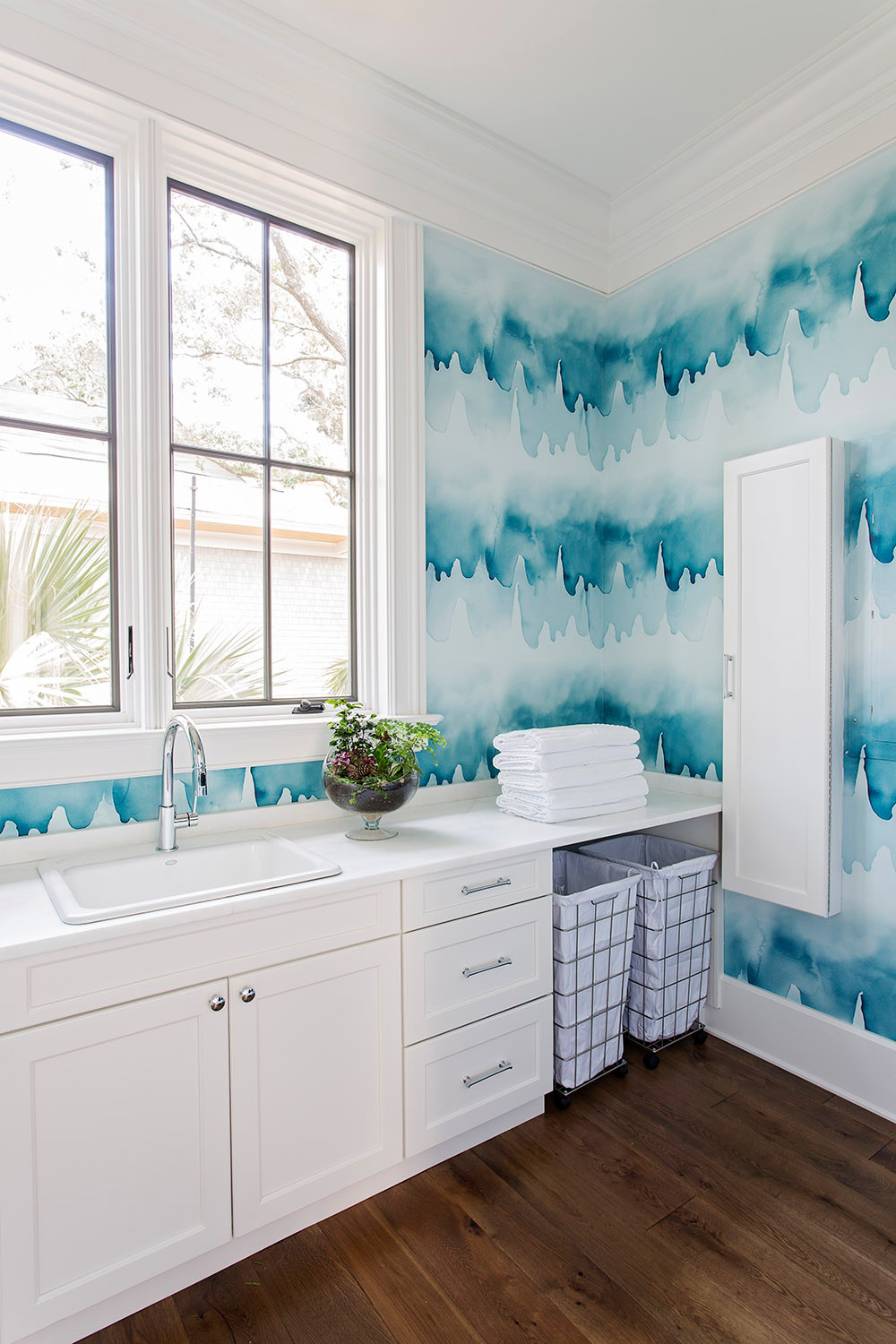Laundry room with aqua watercolor wallpaper, white floating cabinets, chrome faucet, and large windows overlooking palm trees.