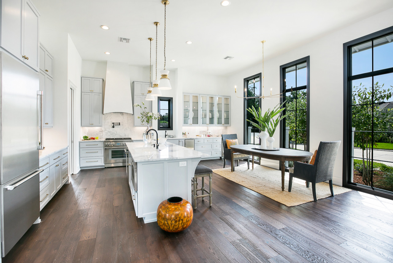 Bright white kitchen with marble counters, light gray cabinets, dark hardwood floors, and black-framed windows overlooking garden.