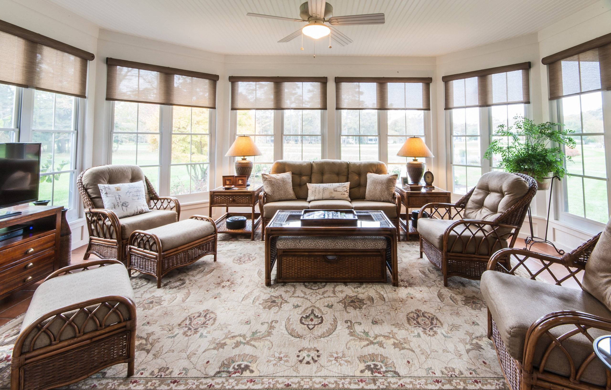 Bright sunroom with rattan furniture set, panoramic windows, linen shades, and floral area rug beneath ceiling fan.