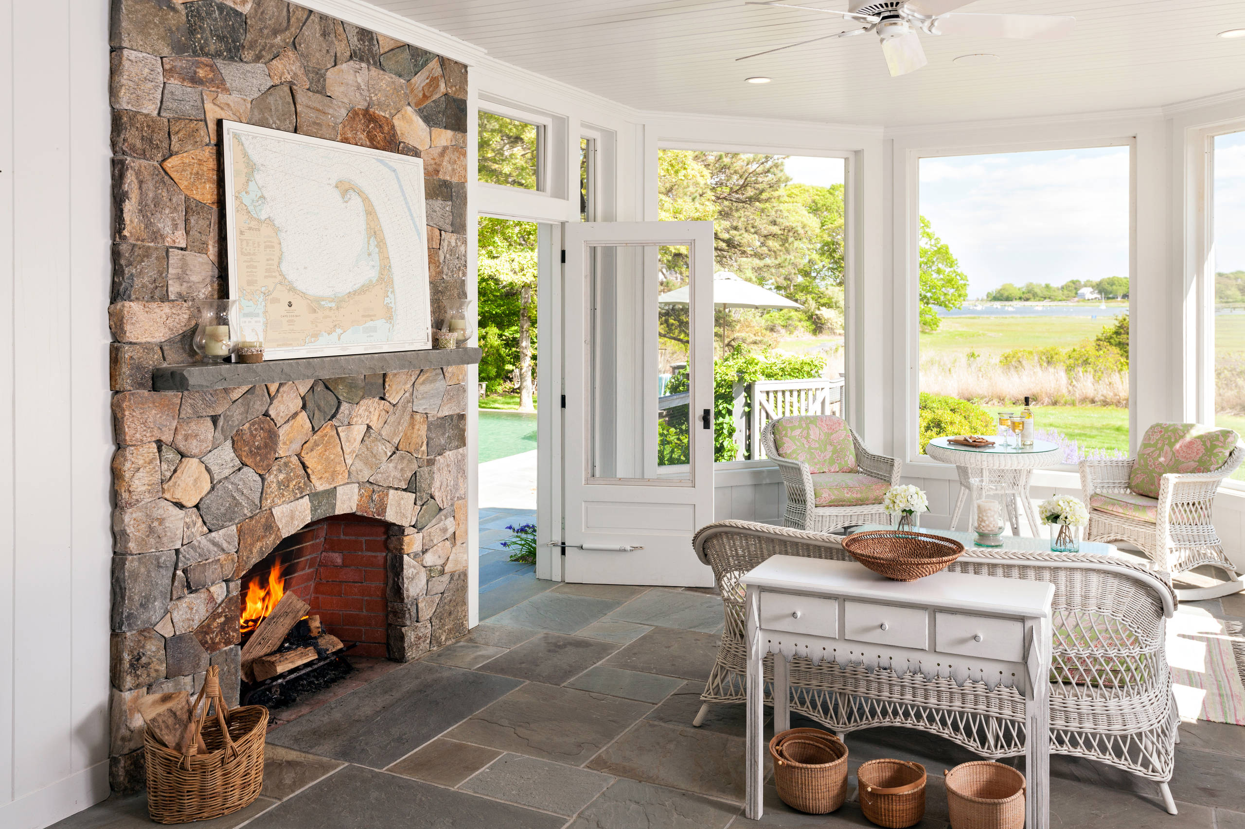 Sunroom with stone fireplace, wicker furniture, slate flooring, and waterfront views through large windows.