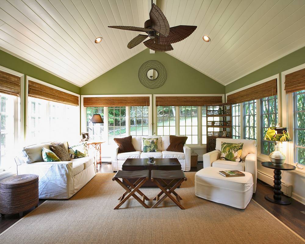 Bright sunroom with vaulted ceiling, white slip-covered furniture, bamboo shades, and sage green walls surrounded by windows.