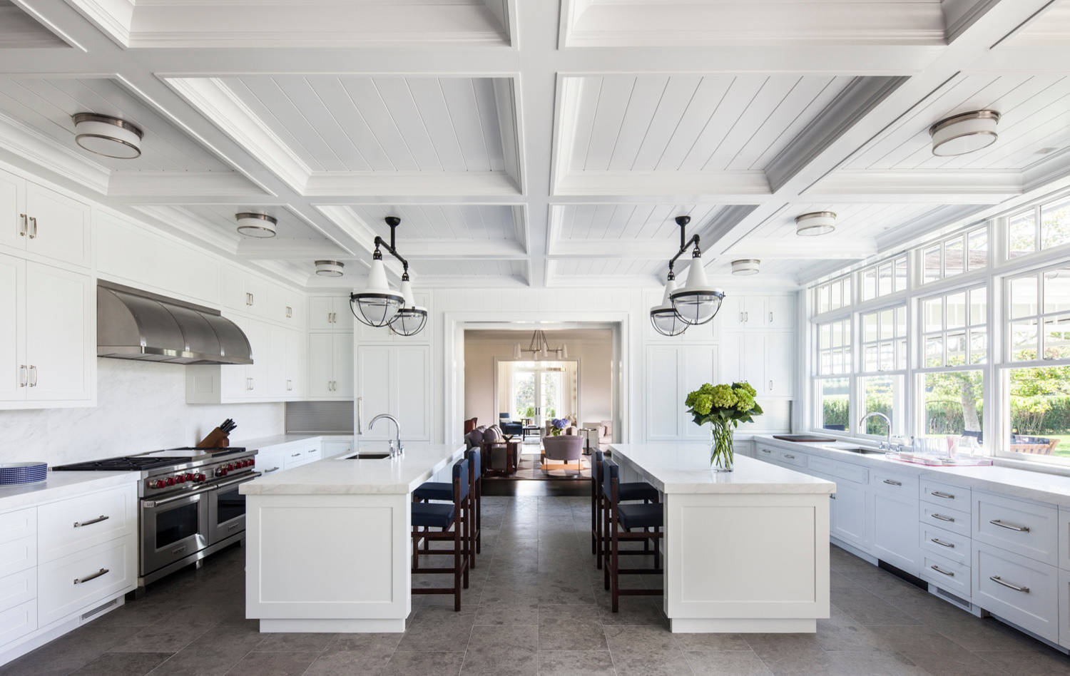 Bright white kitchen with dual islands, coffered ceiling, nautical pendants, and wall of windows featuring professional-grade appliances.