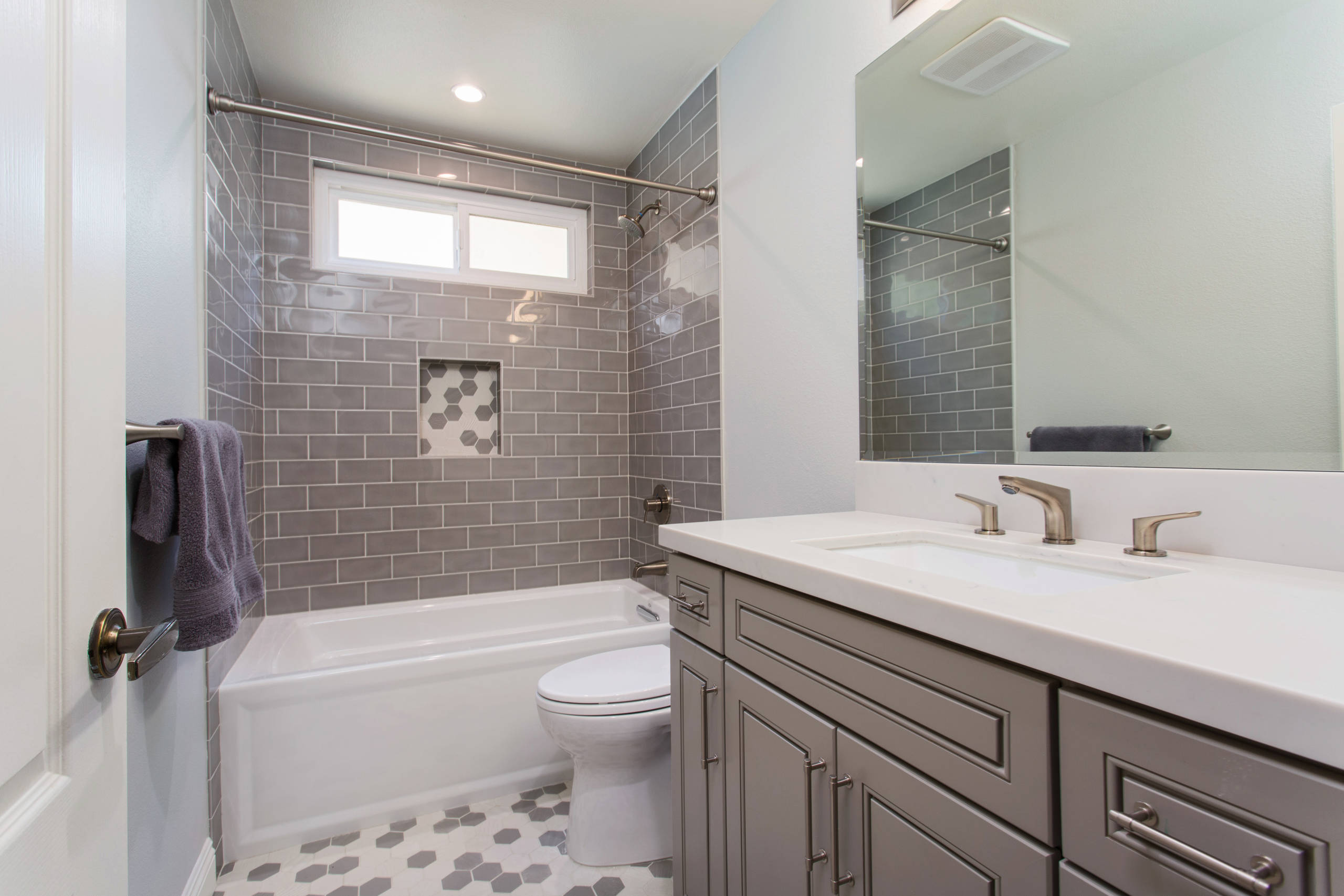 Modern bathroom featuring gray subway tile walls, hexagonal floor tiles, white tub, and taupe vanity with brushed nickel fixtures.