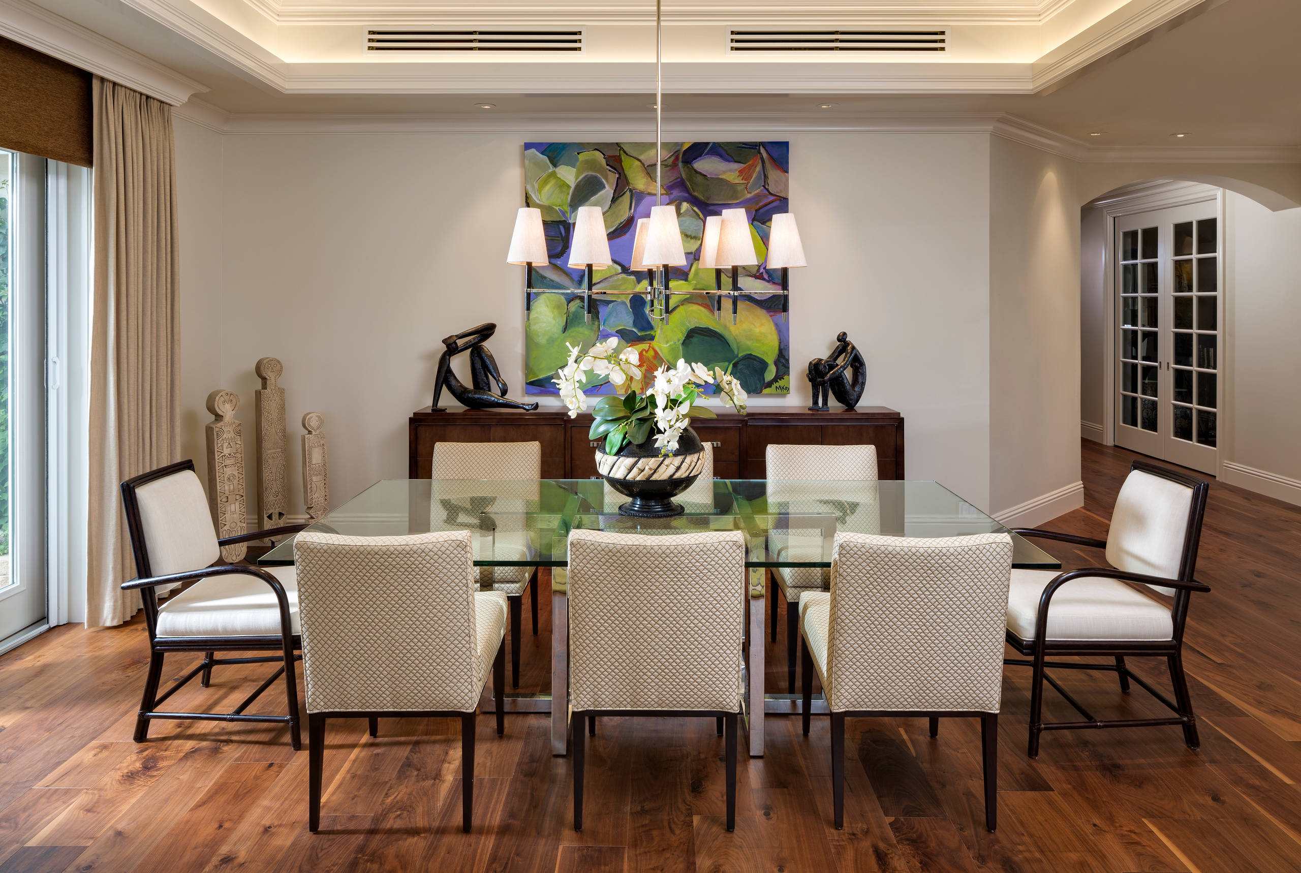 Contemporary dining room featuring glass-top table, cream upholstered chairs, abstract wall art, and sculptural accents on dark wood floors.