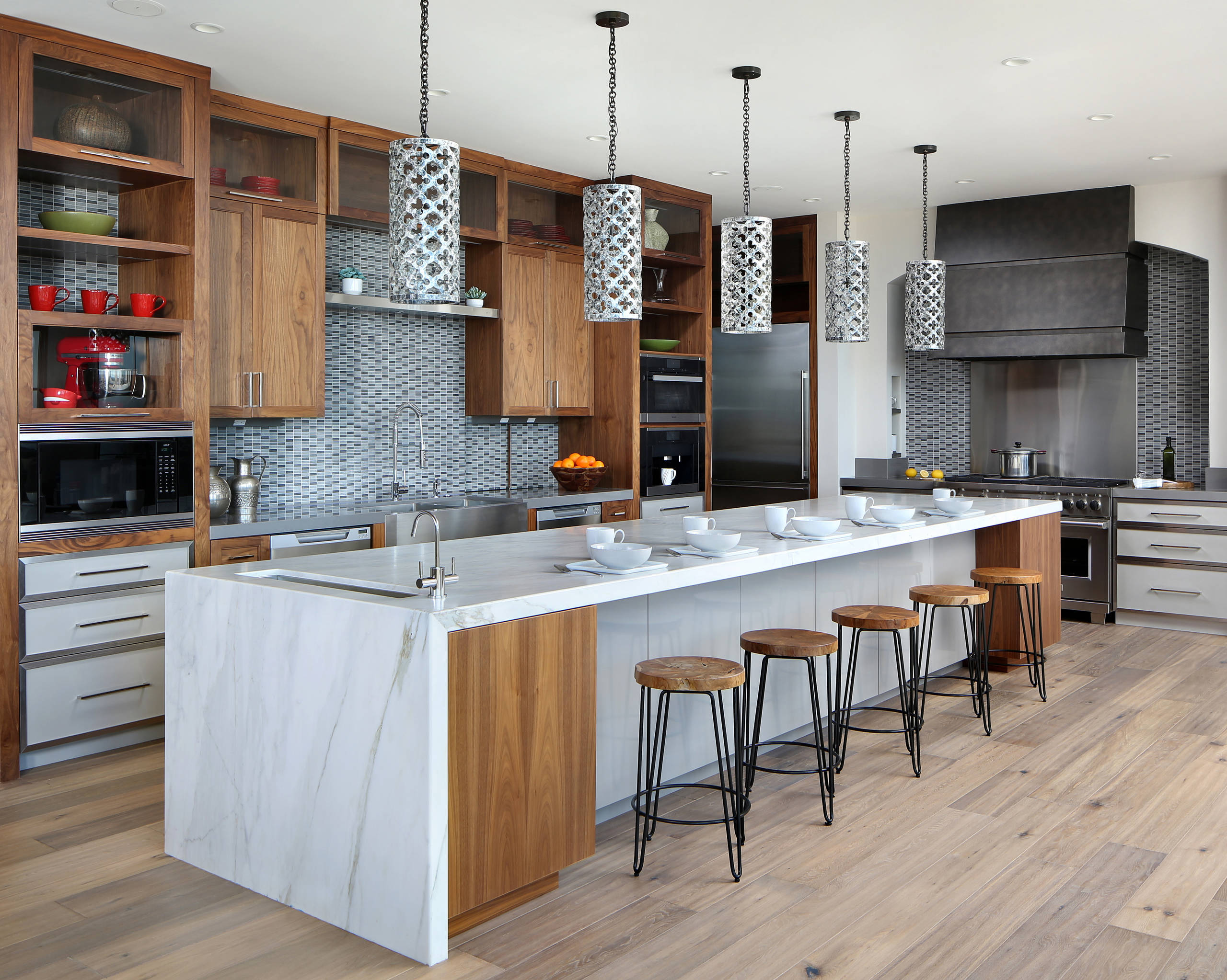 Modern kitchen featuring marble waterfall island, walnut cabinets, patterned pendant lights, and gray tile backsplash with industrial bar stools.