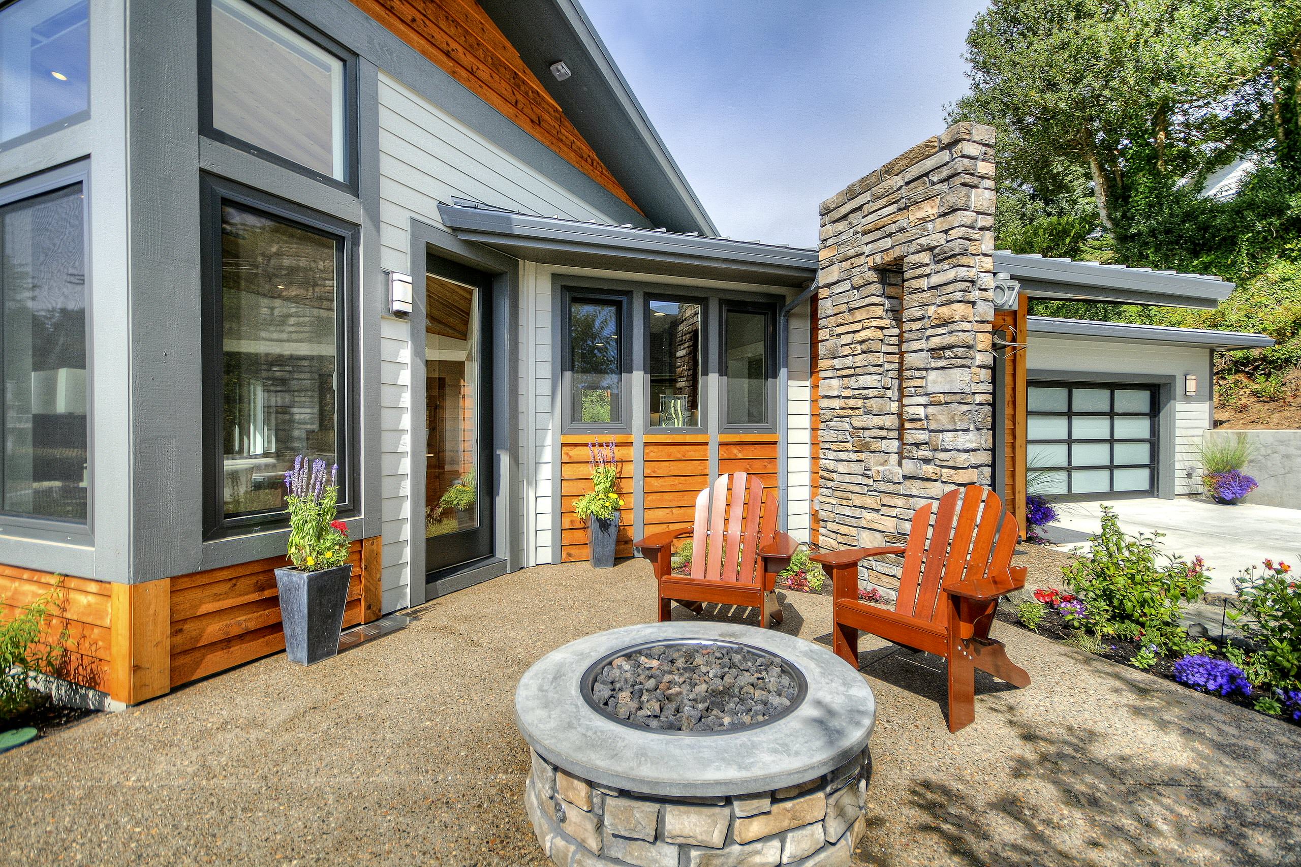 Modern patio featuring circular stone fire pit, cedar accents, Adirondack chairs, and purple flowering plants against contemporary house exterior.