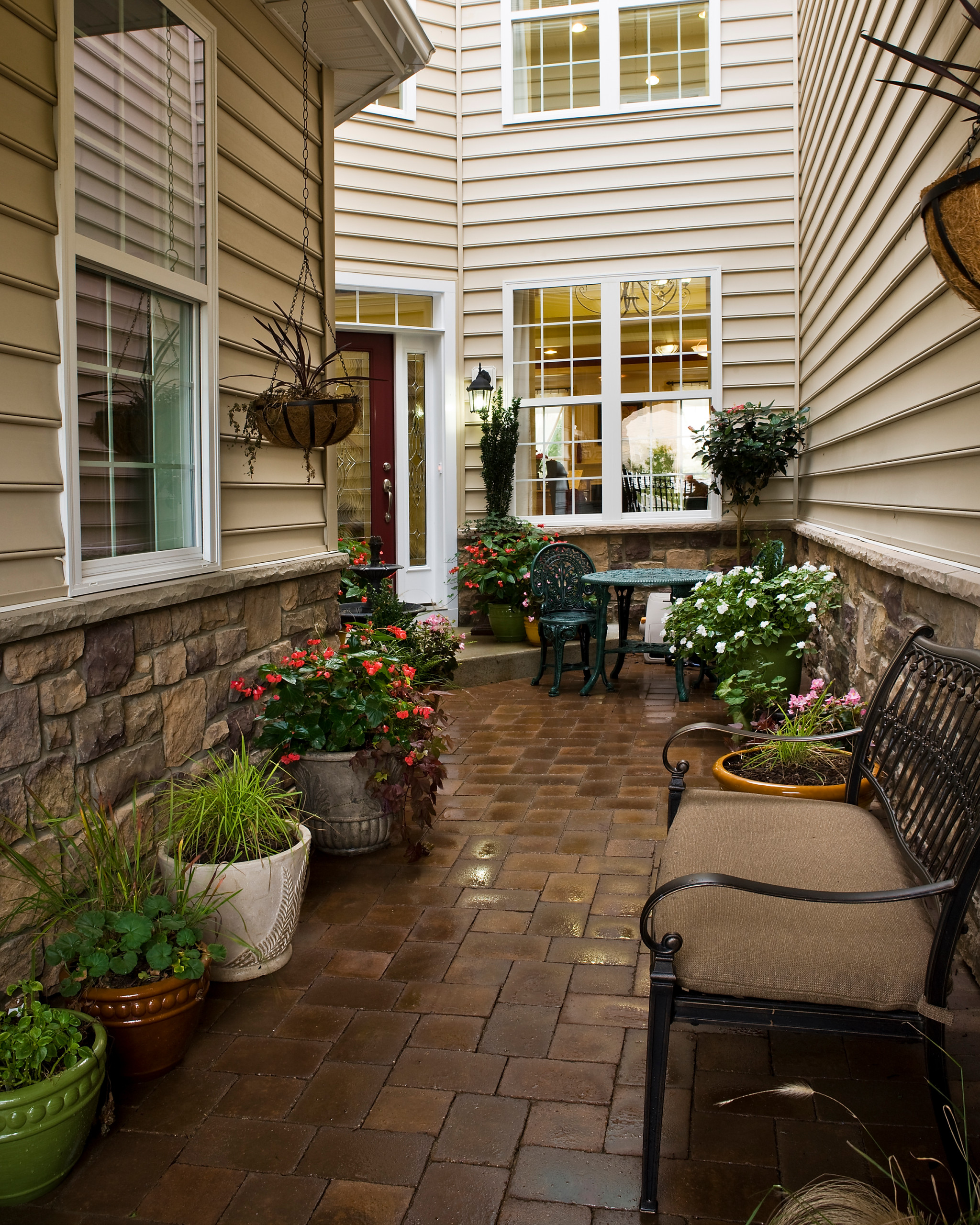 Enclosed courtyard with stone foundation, brick pavers, wrought iron furniture, and colorful container gardens featuring red and white flowers.