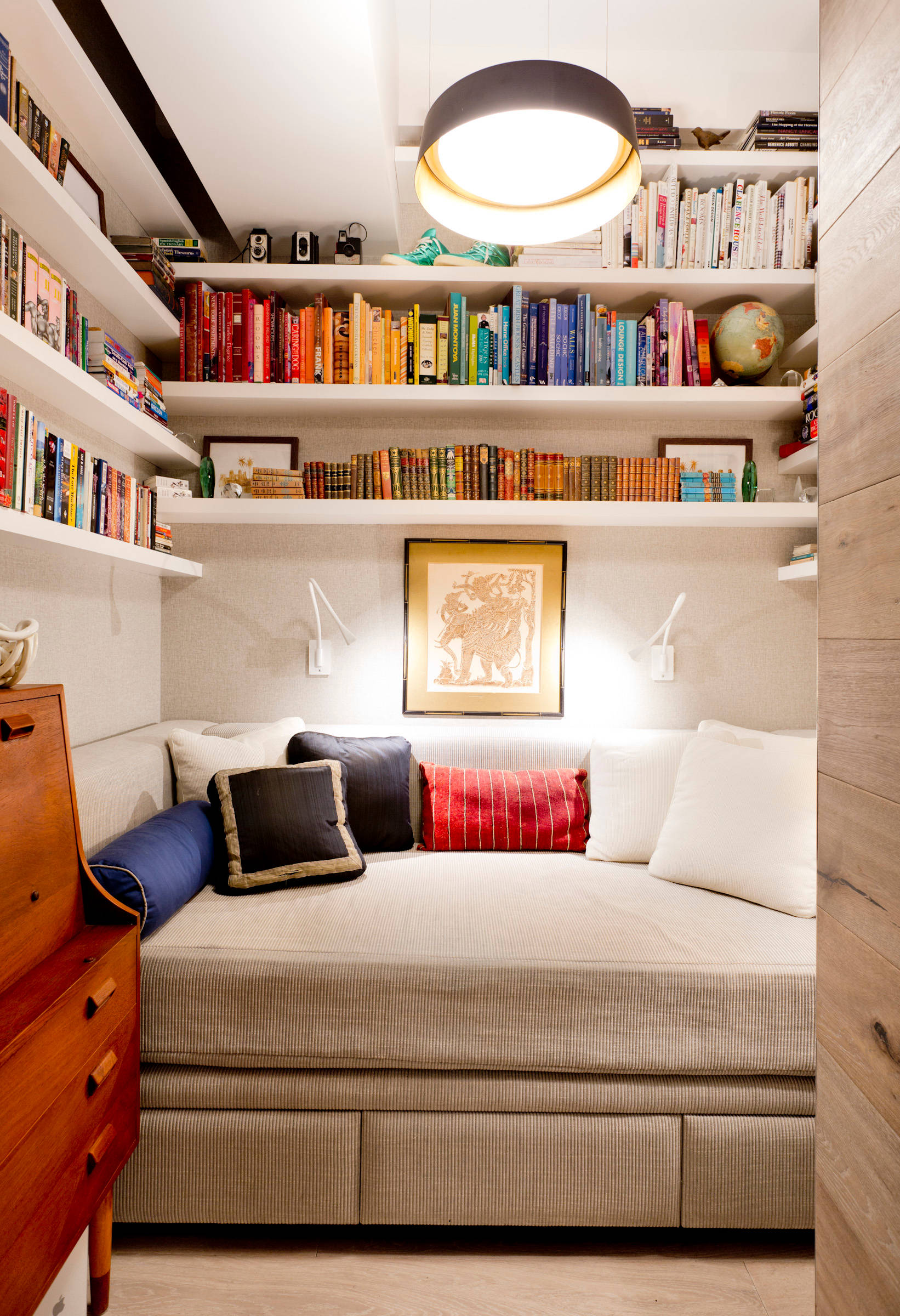 Built-in daybed surrounded by floating white shelves displaying color-coordinated books, with modern pendant light and wooden accents.