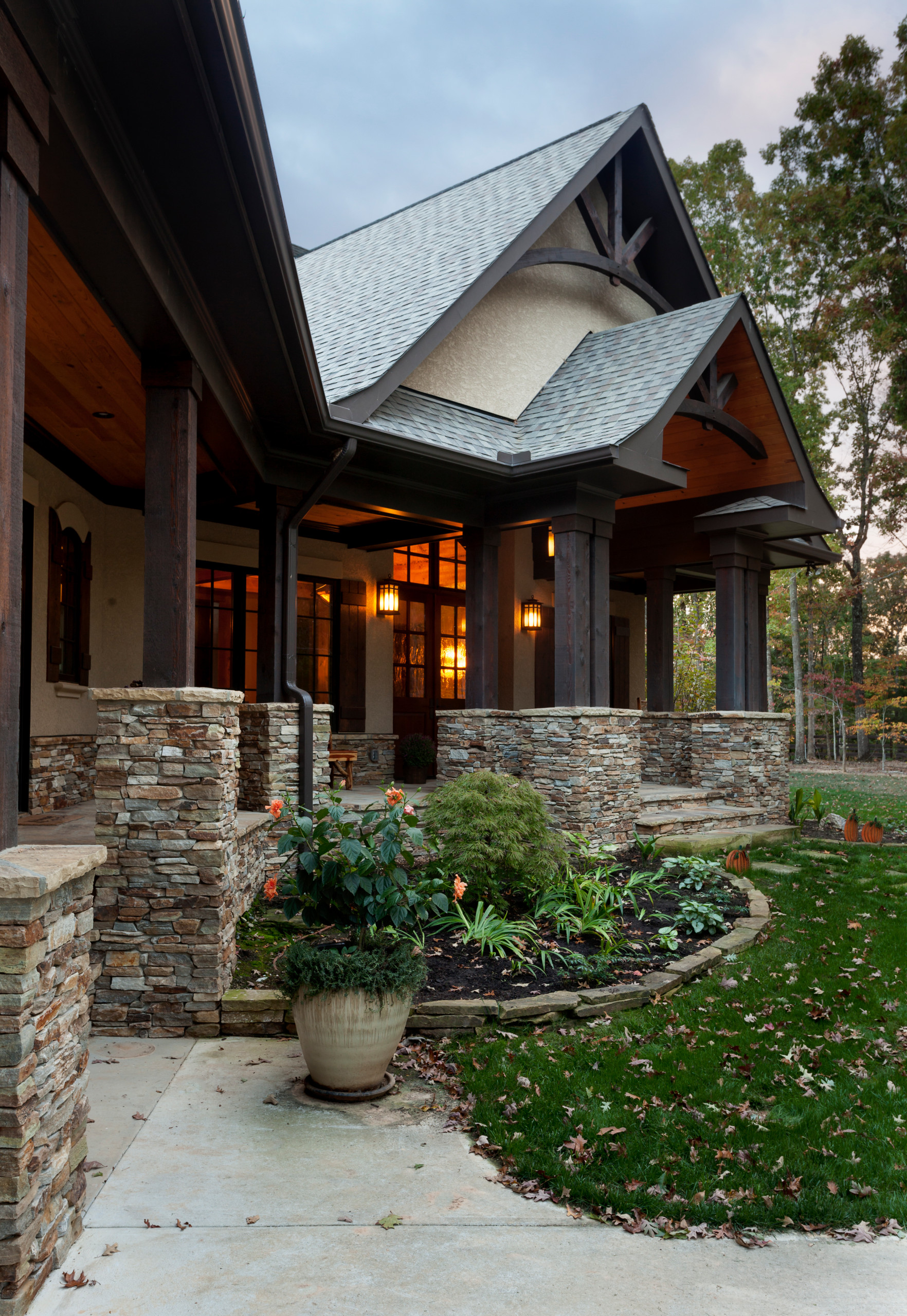 Craftsman-style covered porch with stacked stone pillars, lantern lighting, and landscaped entrance featuring potted plants.