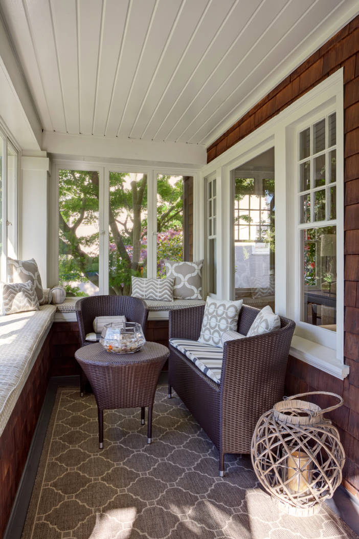 Enclosed porch with dark wicker furniture, geometric patterned pillows, built-in window seats, and white-trimmed windows overlooking garden.