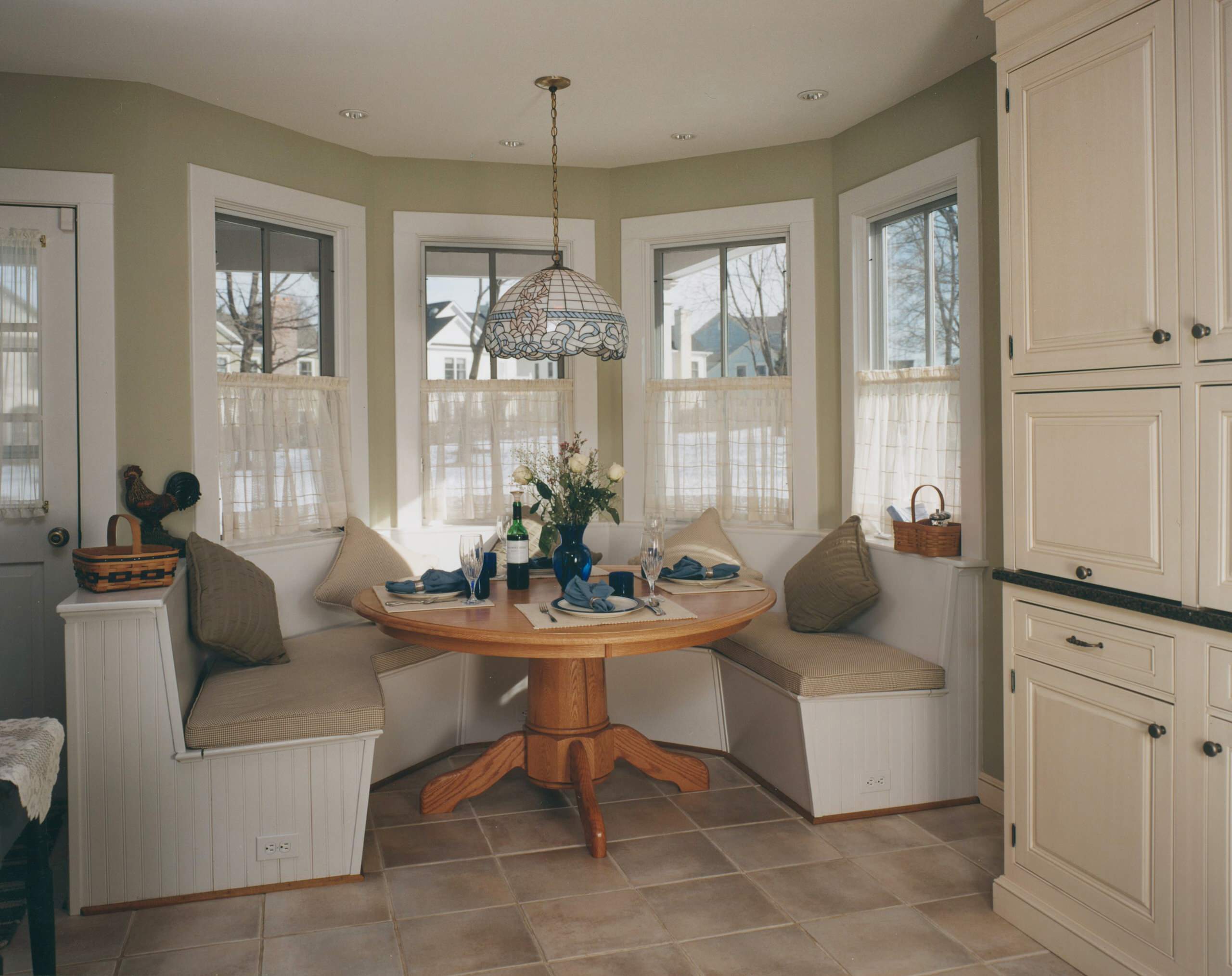 Curved breakfast nook featuring built-in bench seating, oak pedestal table, Tiffany-style pendant light, and cafe curtains in bay window.