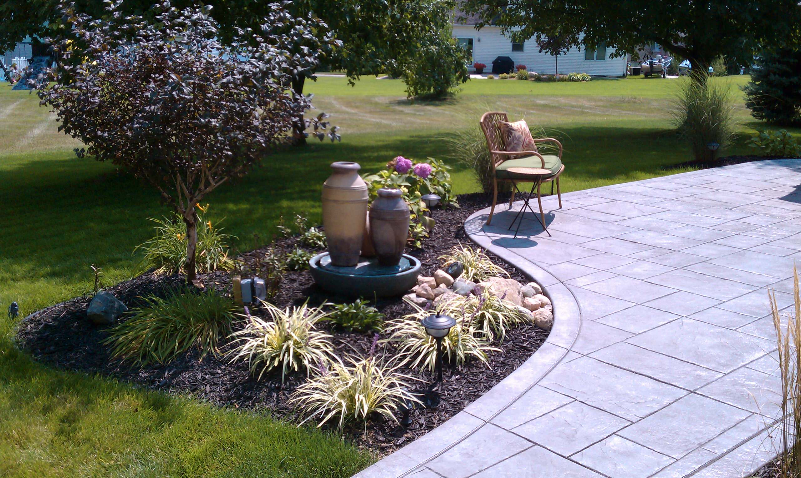 Curved stone patio with ceramic fountain feature, ornamental grasses, and wicker chair beside landscaped garden bed.