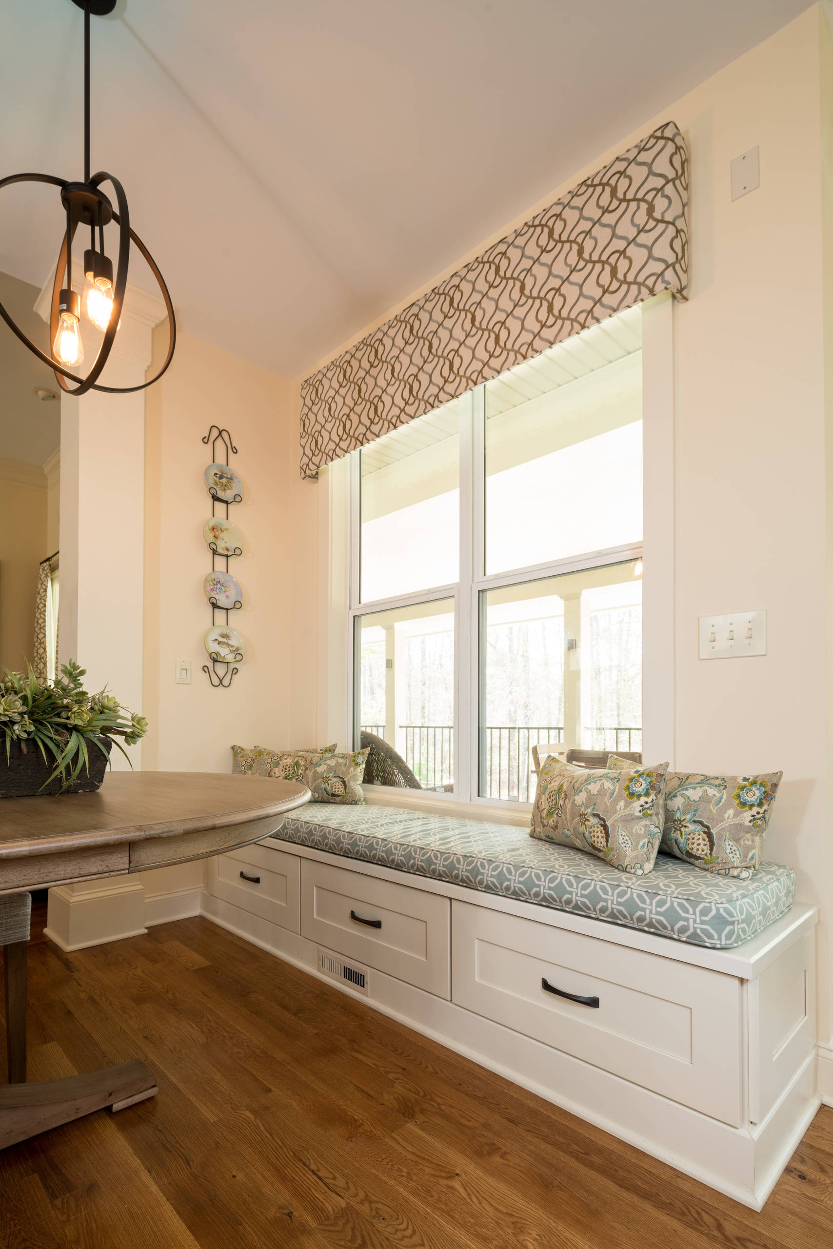 Built-in white banquette with storage drawers beneath large window, circular dining table, and geometric valance.