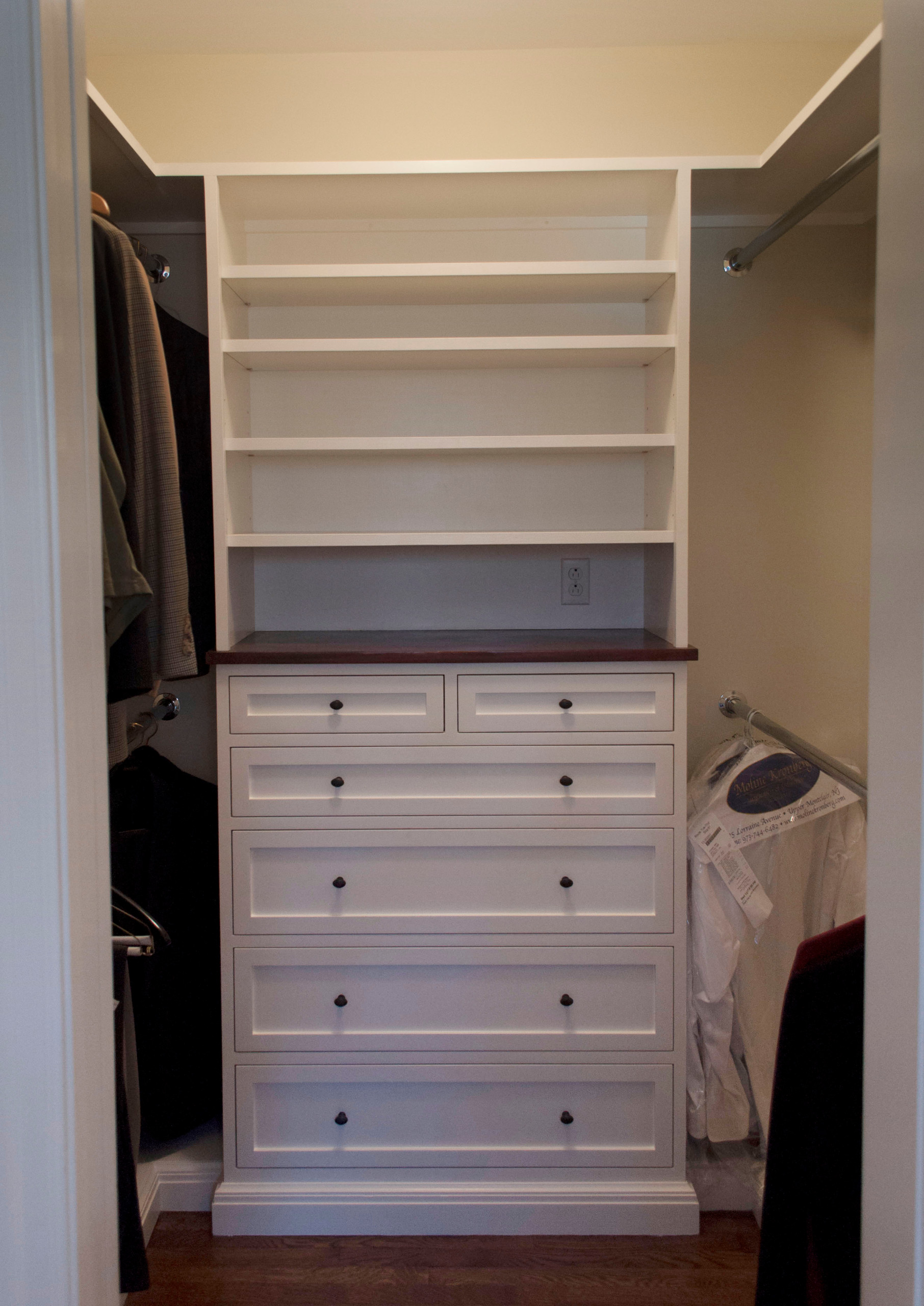 Walk-in closet featuring white built-in drawers with black hardware, open shelving, walnut countertop, and hanging rods on both sides.