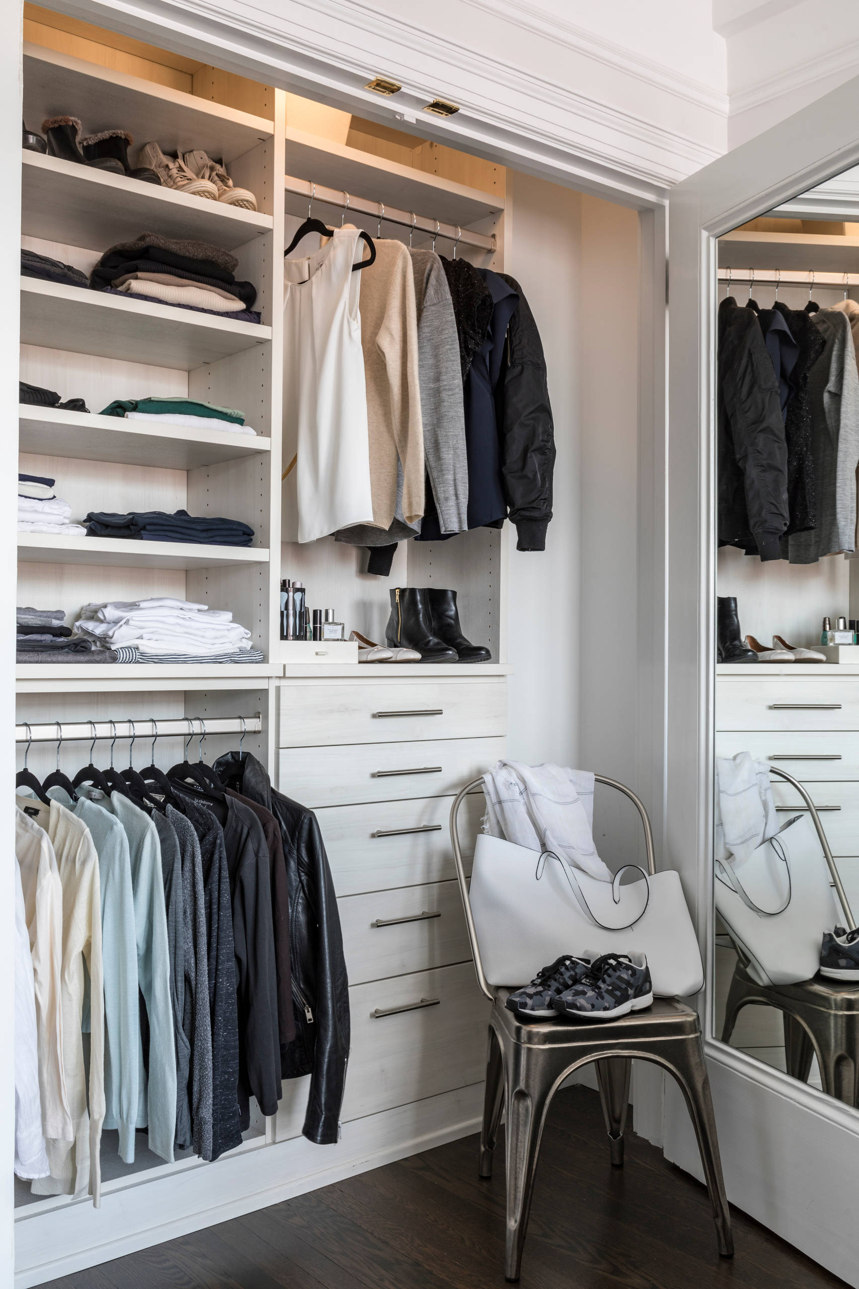 Built-in white closet with organized clothing racks, open shelving, drawers, metal chair, and full-length mirror on dark wood flooring.