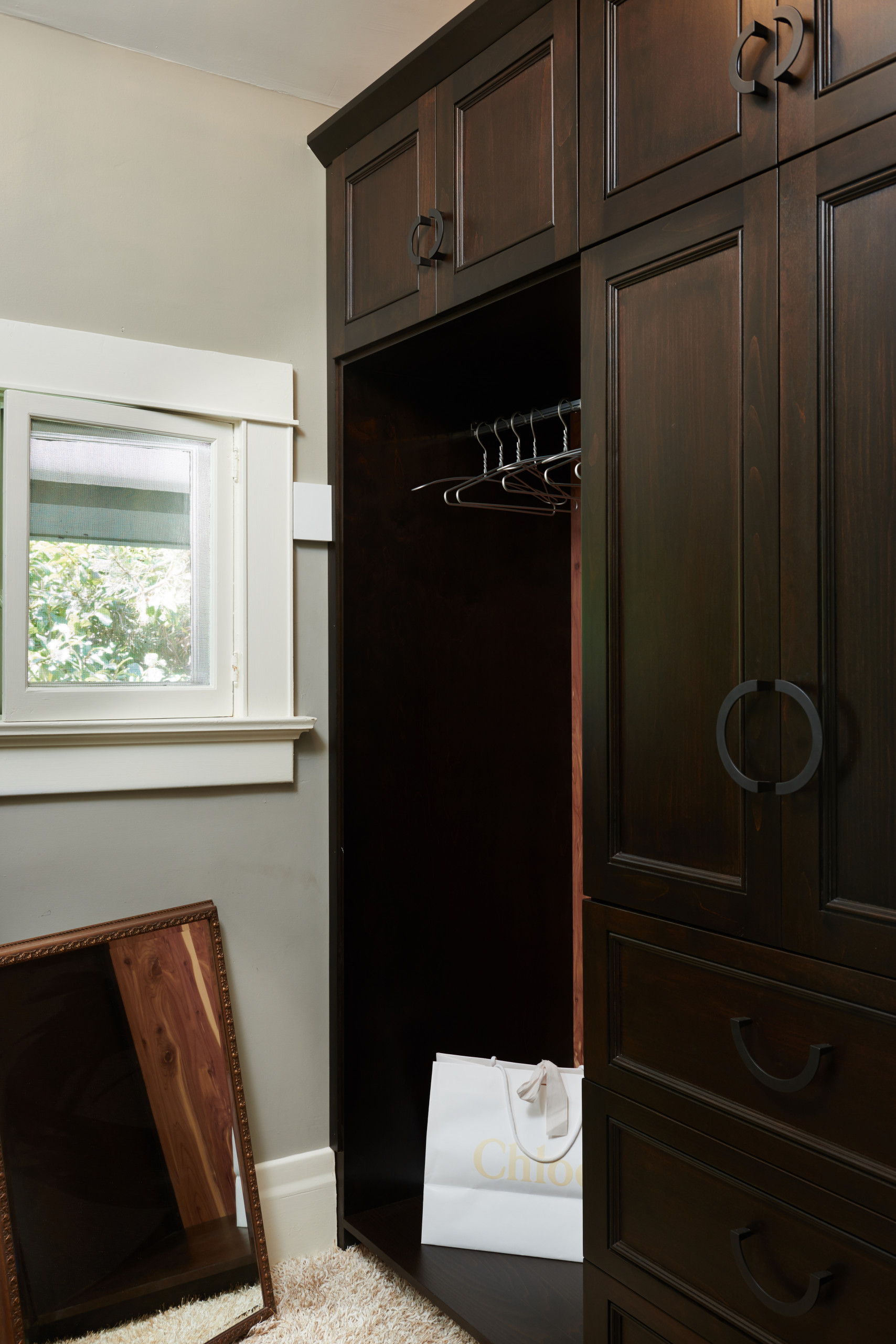 Rich mahogany built-in wardrobe with circular hardware, open hanging space, small window, and cream walls in dressing area.