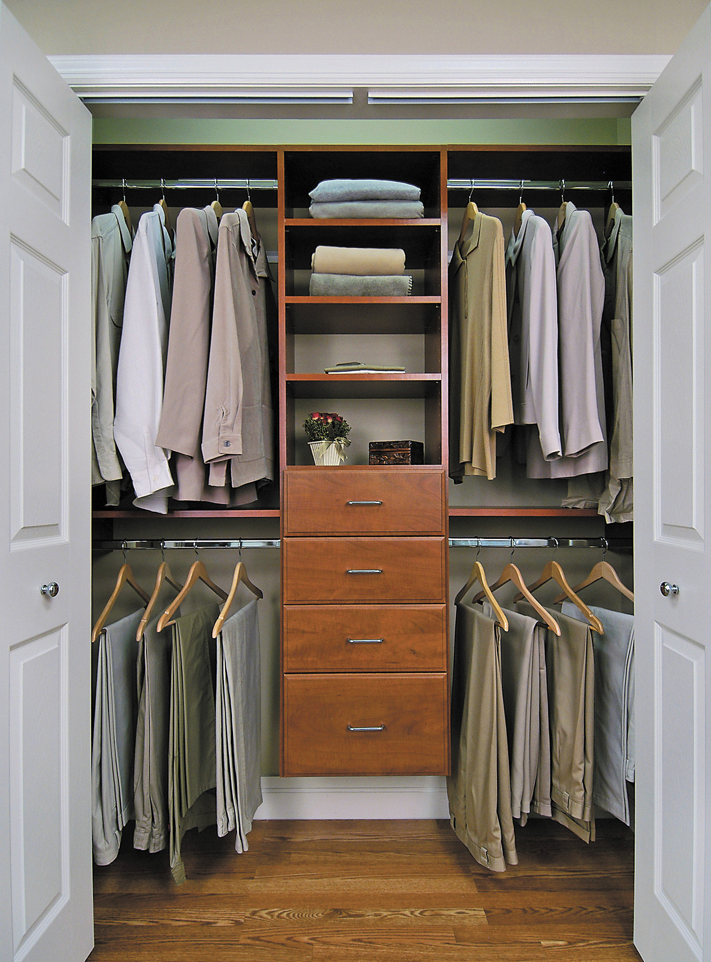 Organized reach-in closet with cherry wood cabinetry, hanging rods for shirts and pants, and central drawers with open shelving above.