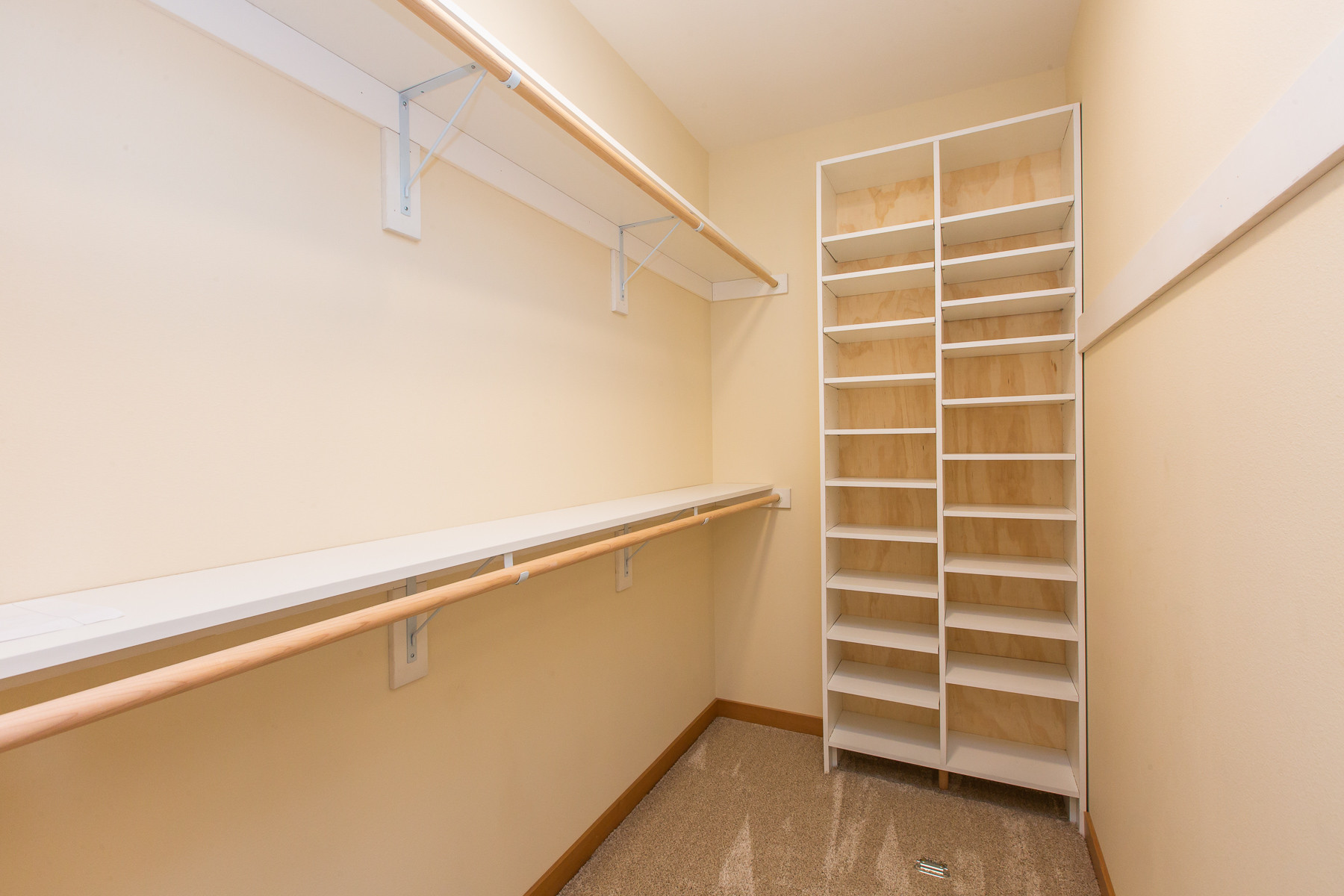 Walk-in closet featuring white built-in shelving unit with wood backing, hanging rods, and neutral cream walls with beige carpeting.