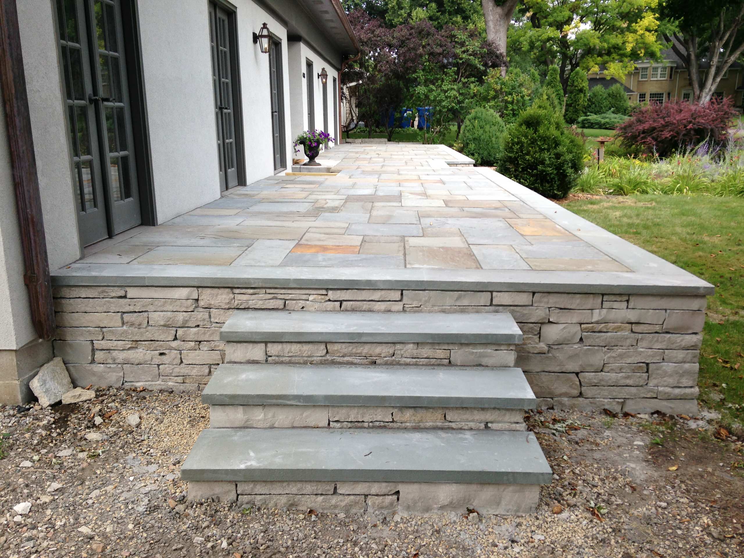 Elevated porch with variegated bluestone pavers, stacked stone foundation, and concrete steps leading to landscaped garden.