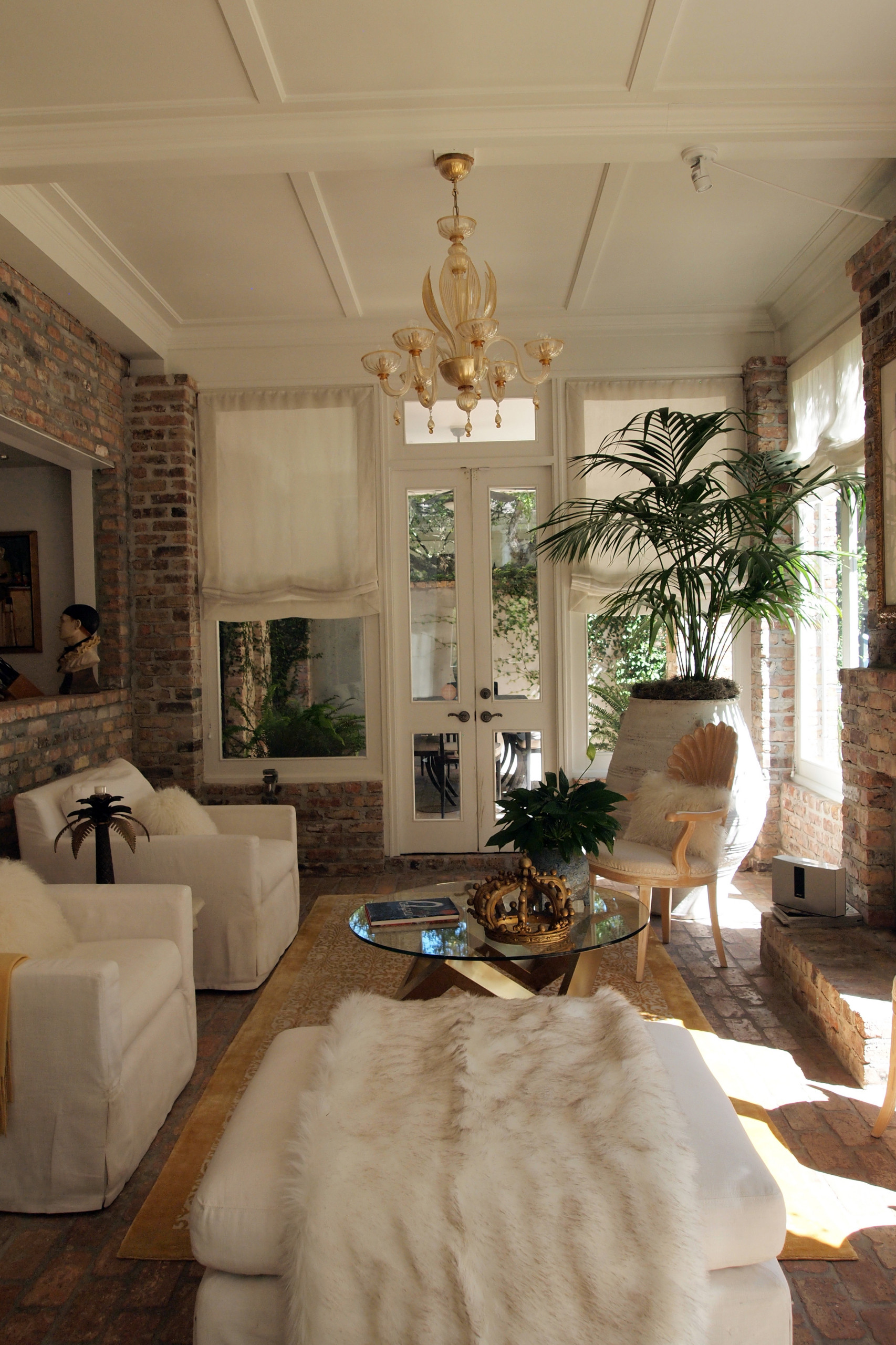 Sunlit living space with exposed brick walls, white slipcovered furniture, amber chandelier, and tropical plants against French doors.