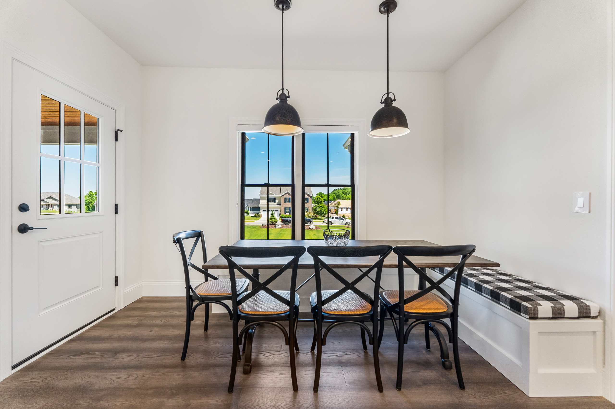 Bright breakfast nook with black crossback chairs, industrial pendants, built-in bench, and large windows overlooking suburban view.