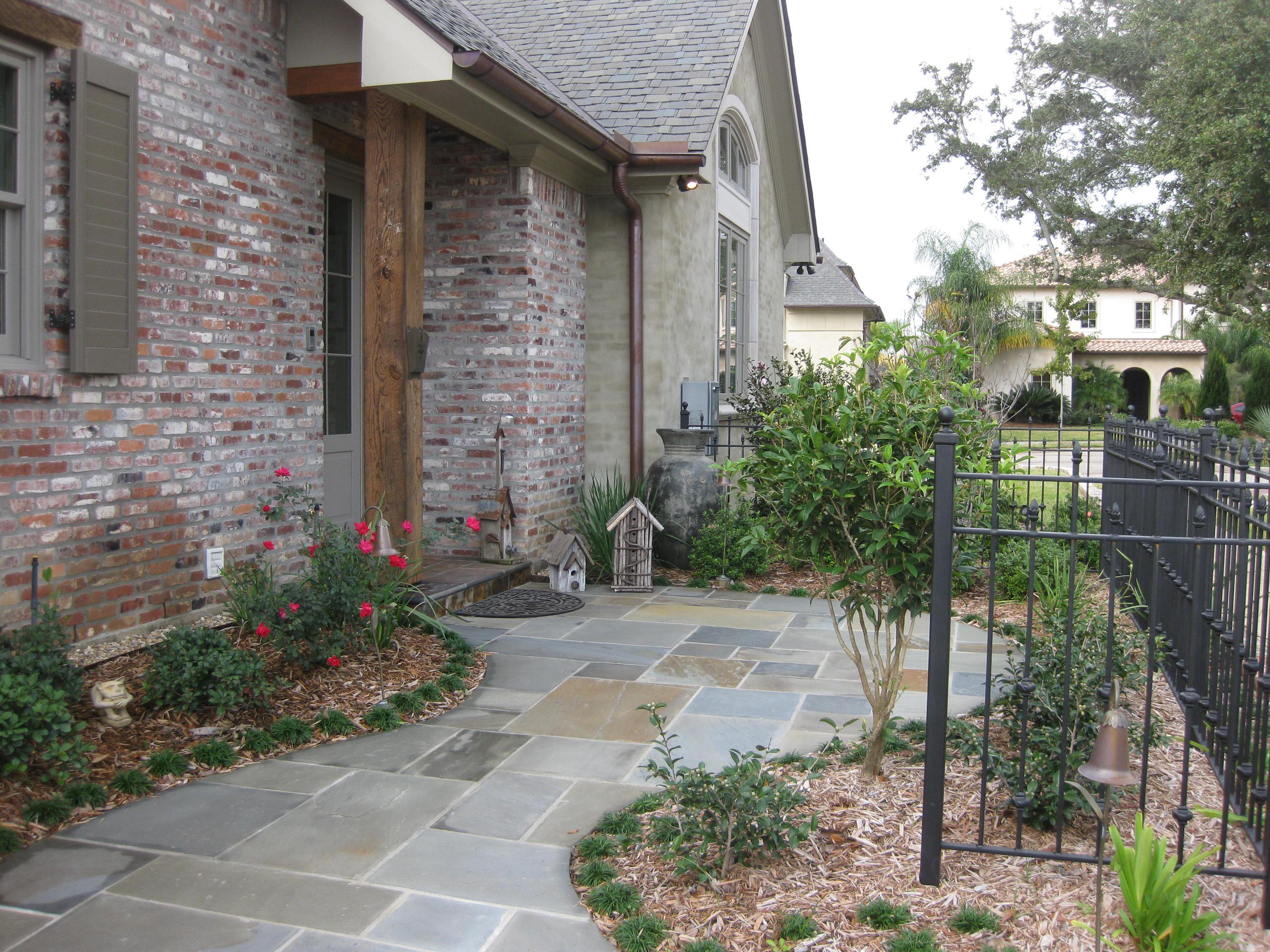 Bluestone pathway leading to brick home entrance, featuring cottage garden plantings, wrought iron fence, and decorative birdhouses.
