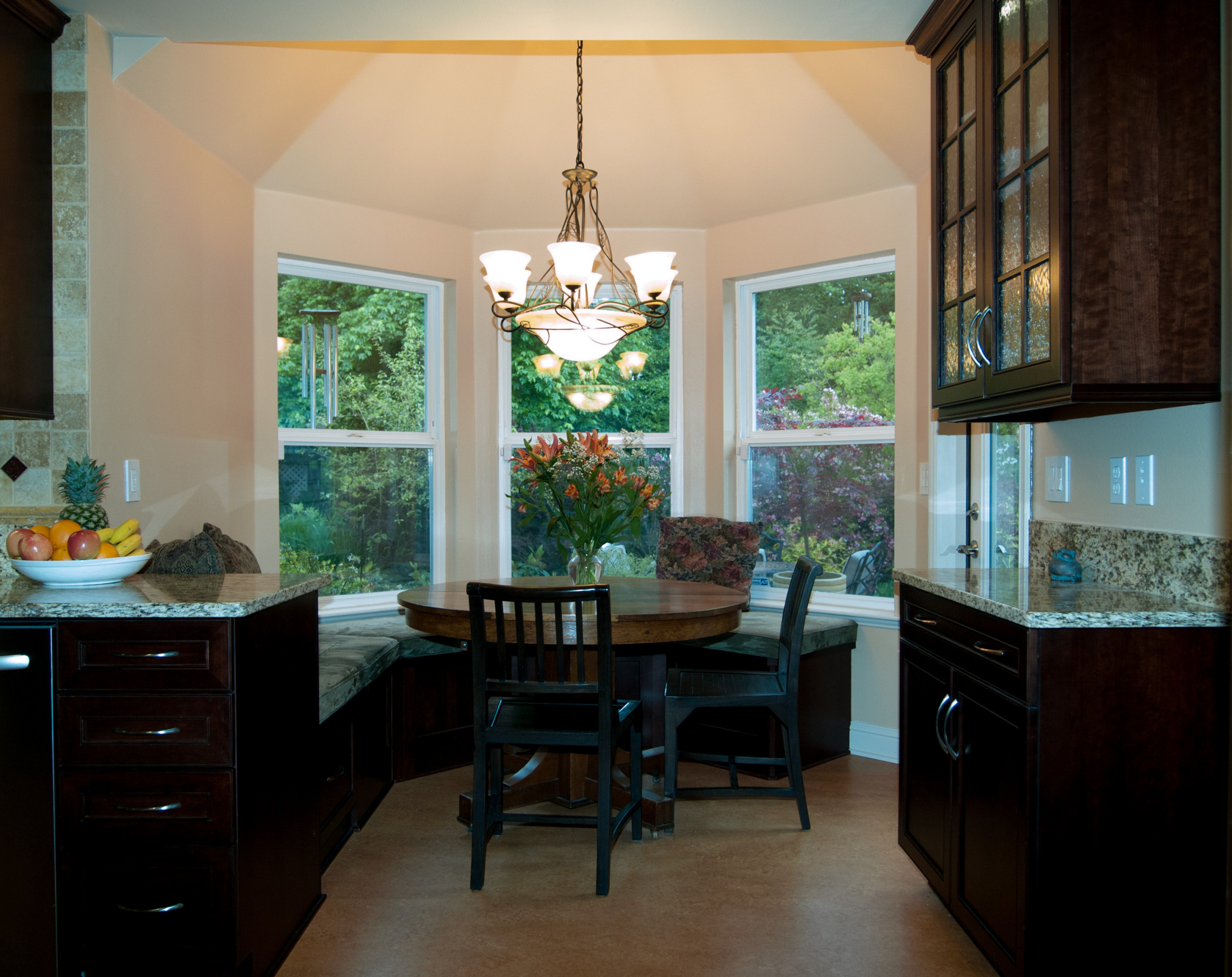 Breakfast nook with bay windows, round wooden table, black chairs, dark cabinets, and granite countertops illuminated by chandelier.
