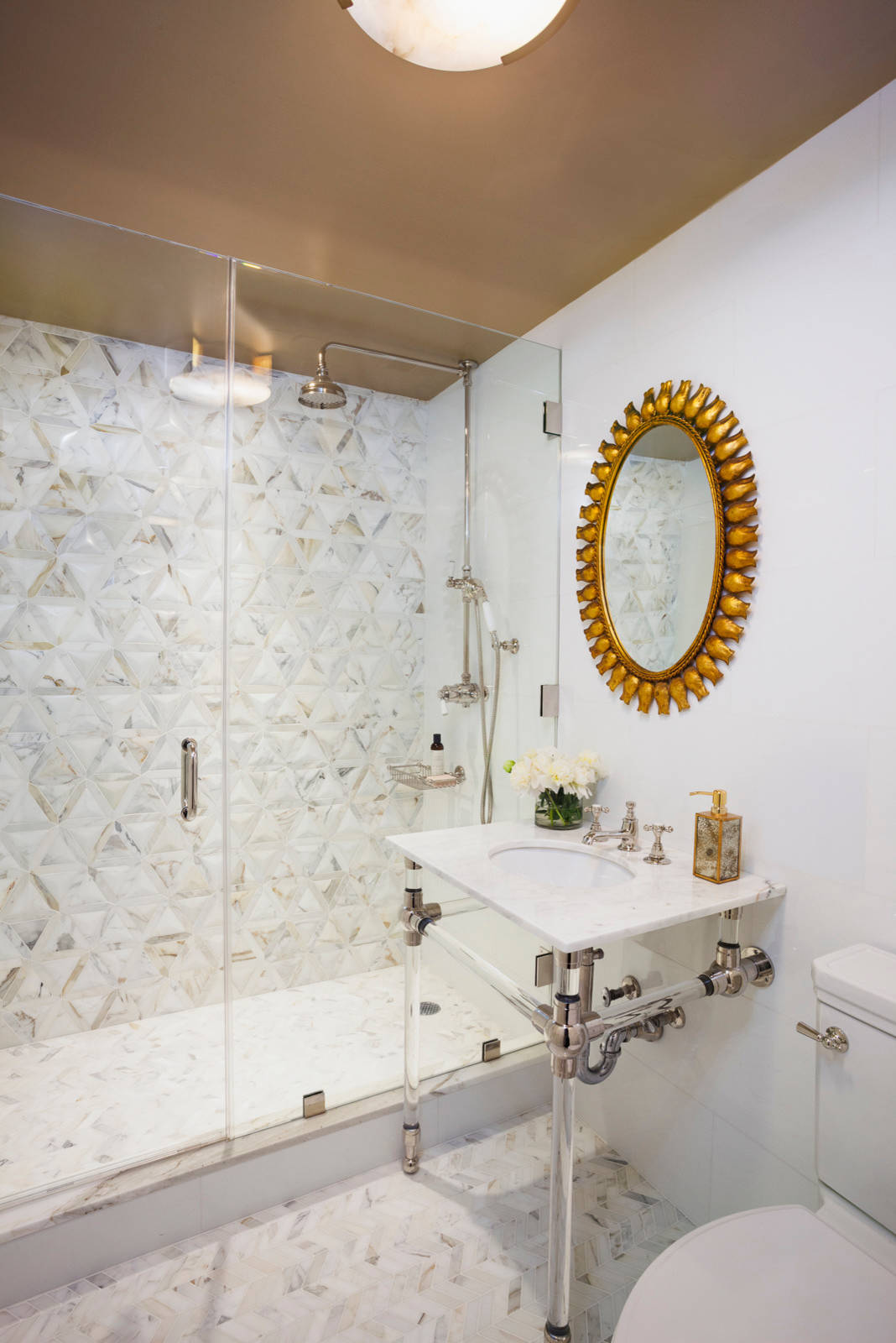 Modern bathroom featuring geometric marble tile walls, glass shower enclosure, white console sink, and gold sunburst mirror.