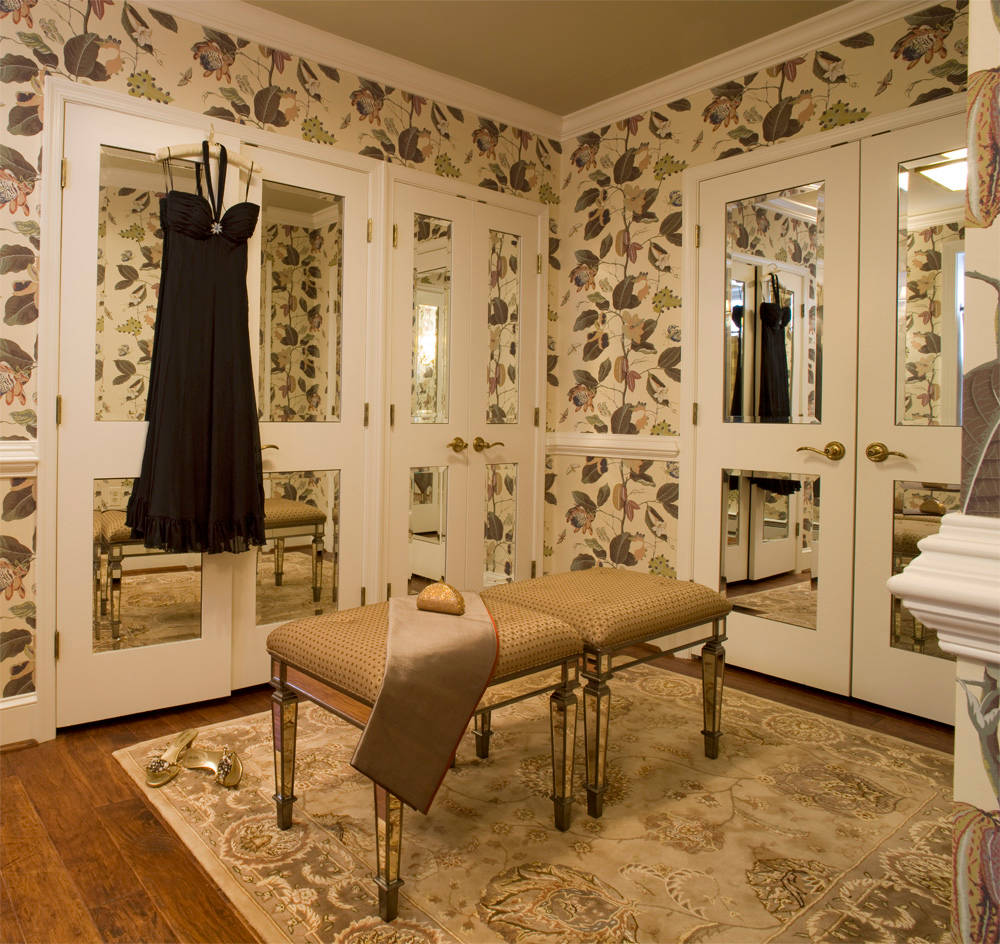 Dressing room featuring floral wallpaper, mirrored closet doors, gold-legged benches, and an ornate area rug on hardwood floors.