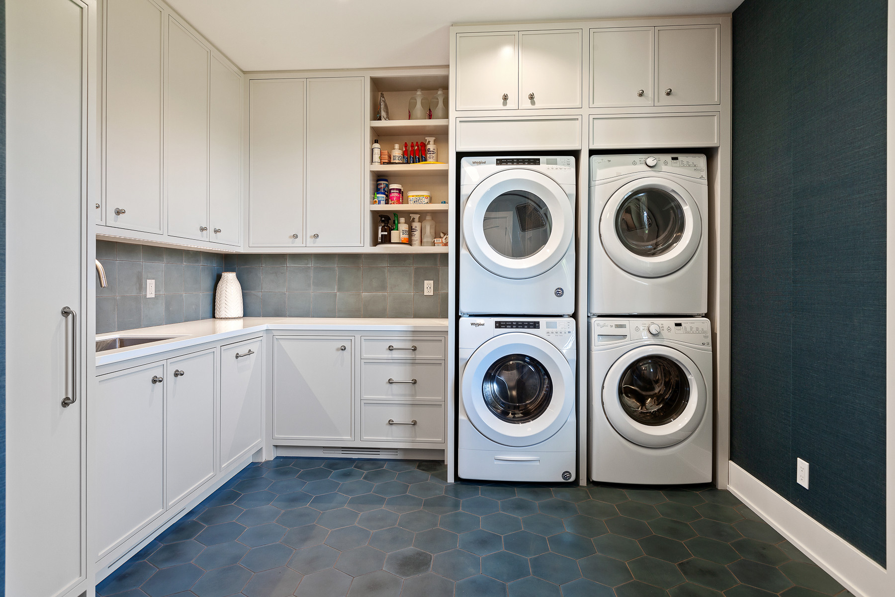 L-shaped laundry room with double stacked washers and dryers, white cabinetry, blue hexagonal floor tiles, and gray backsplash.