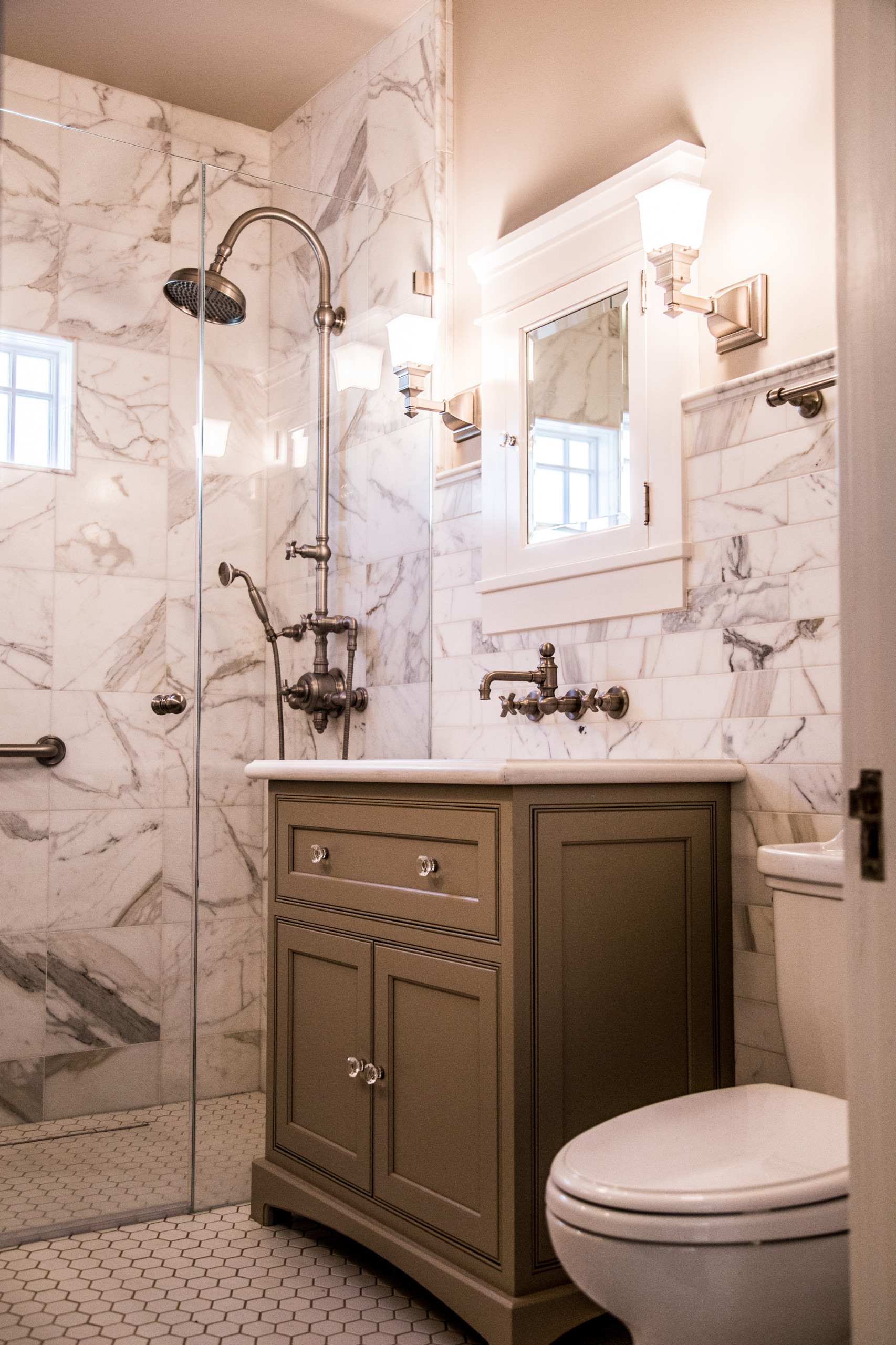 Bathroom featuring marble walls, glass shower enclosure, taupe vanity, bronze fixtures, and hexagonal floor tiles.