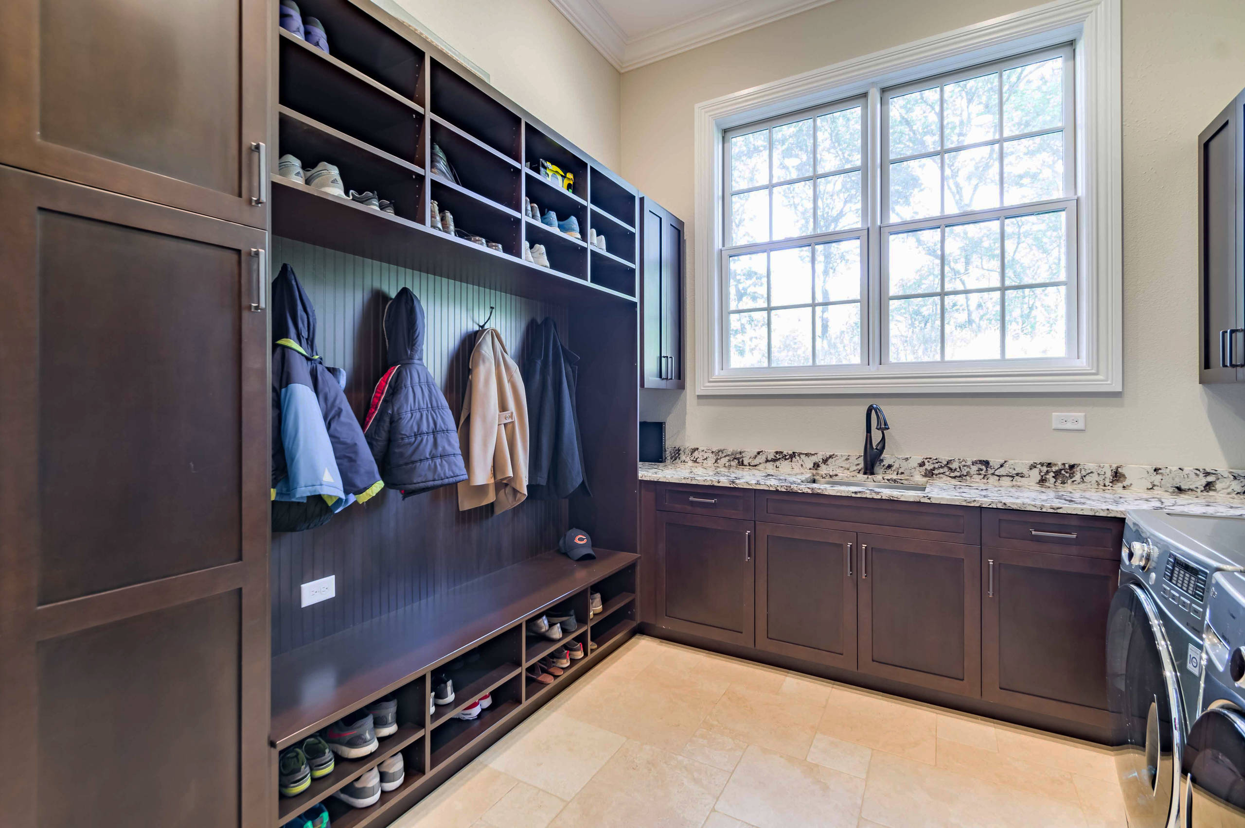 L-shaped mudroom featuring dark wood built-ins, granite countertops, shoe storage, coat hooks, and large window above washer/dryer.