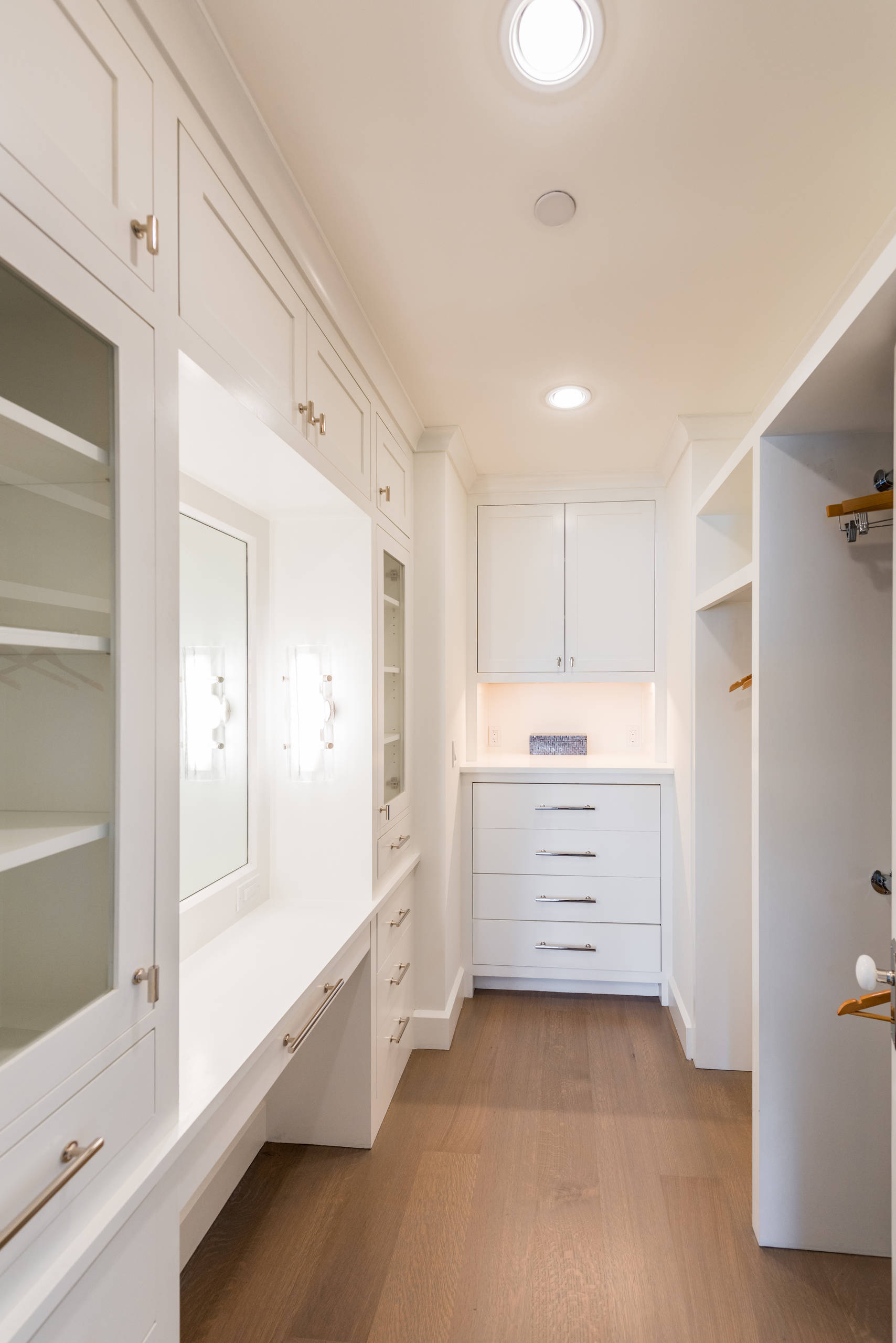 Walk-in closet with crisp white cabinetry, mirrored vanity area, circular skylight, and warm oak flooring throughout.