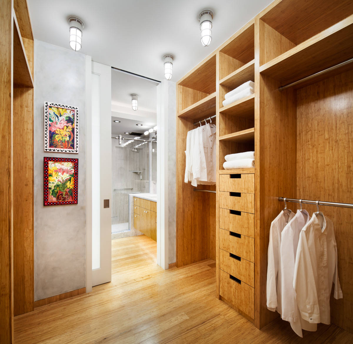 Walk-in closet with honey-toned wood cabinetry, vibrant artwork, light oak flooring, and glimpse into adjacent bathroom.