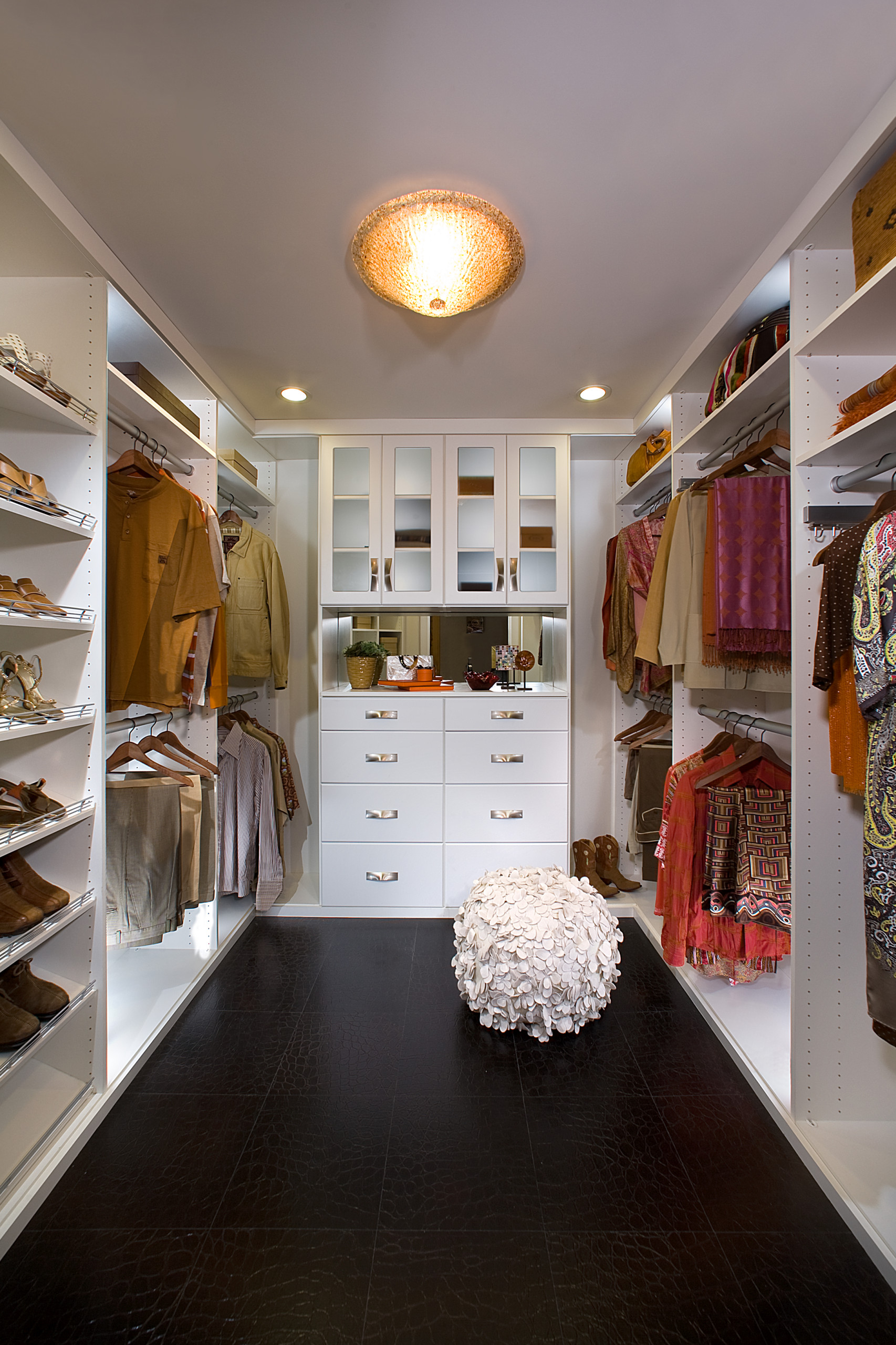 Walk-in closet with white built-in storage, glass cabinet doors, central dresser, dark floors, and decorative textured pouf.