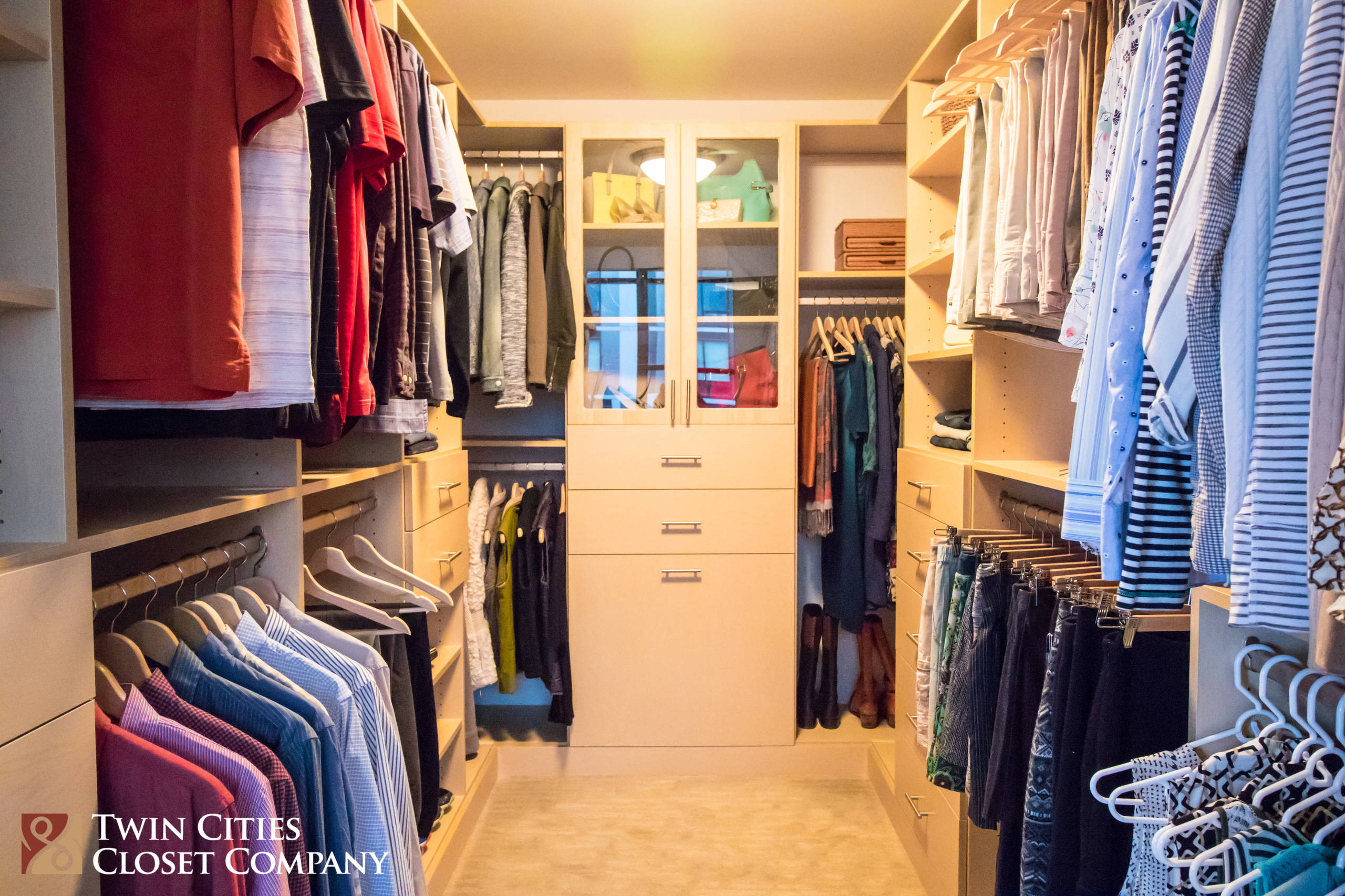 Walkthrough closet with light wood cabinetry, glass-front display case, hanging clothes sections, and organized drawers creating efficient storage.