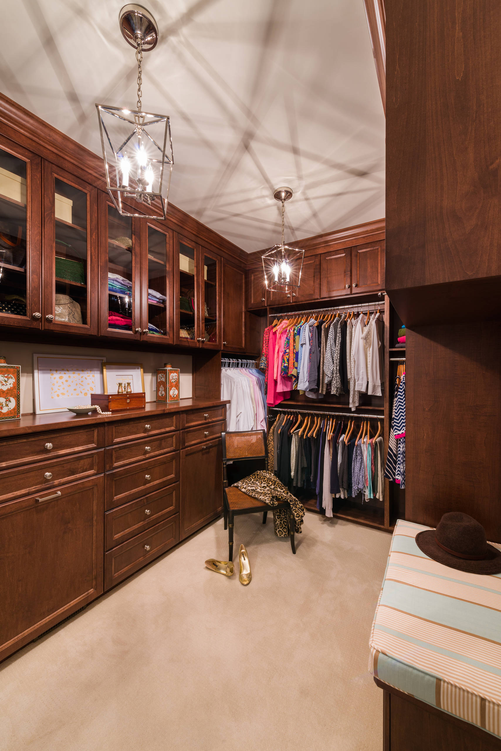 Rich mahogany walk-in closet with glass-front cabinets, geometric pendant lighting, built-in drawers, and organized clothing racks.